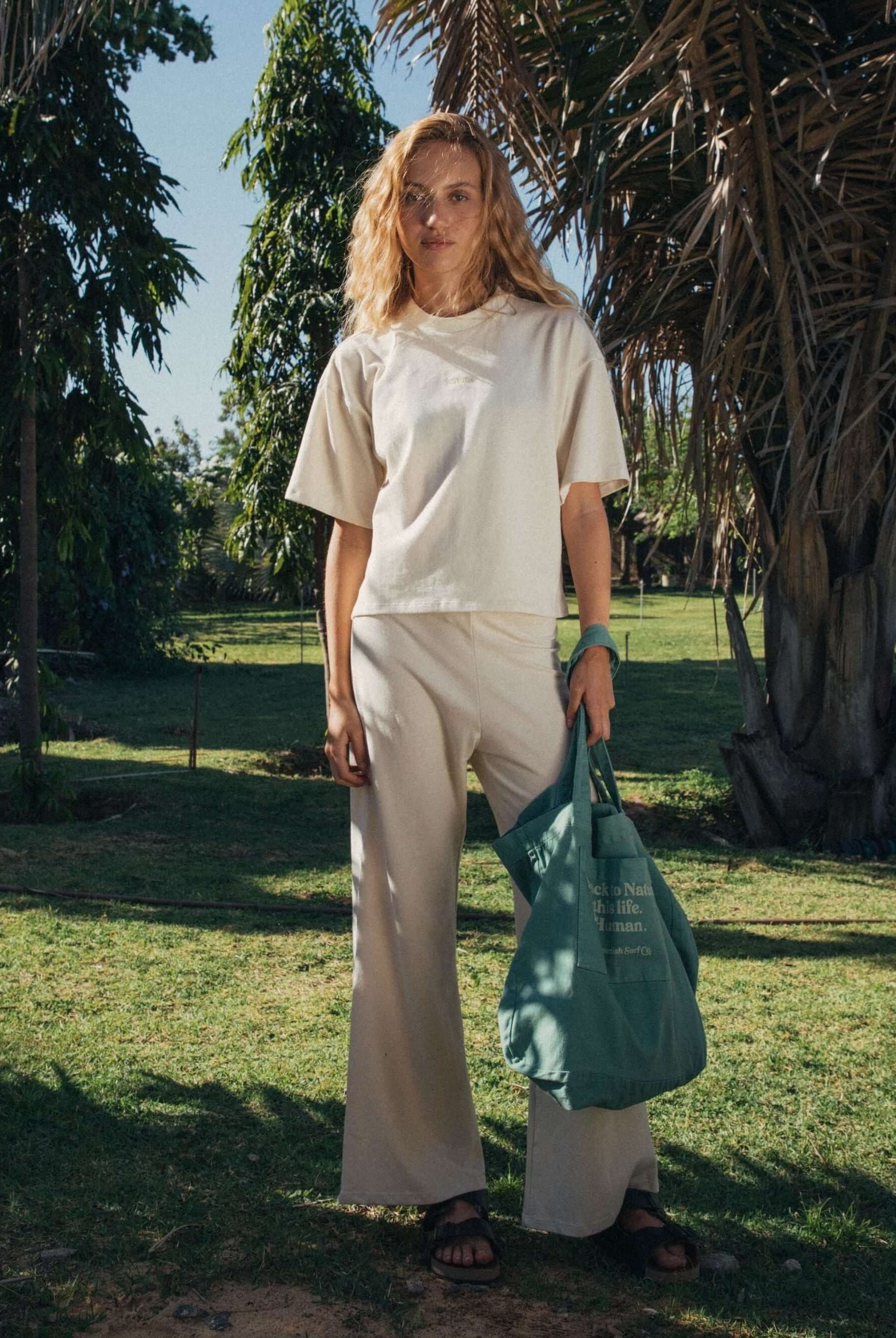 Modelo usando calça Gina bege da Greenish, em ambiente natural com palmeiras ao fundo.