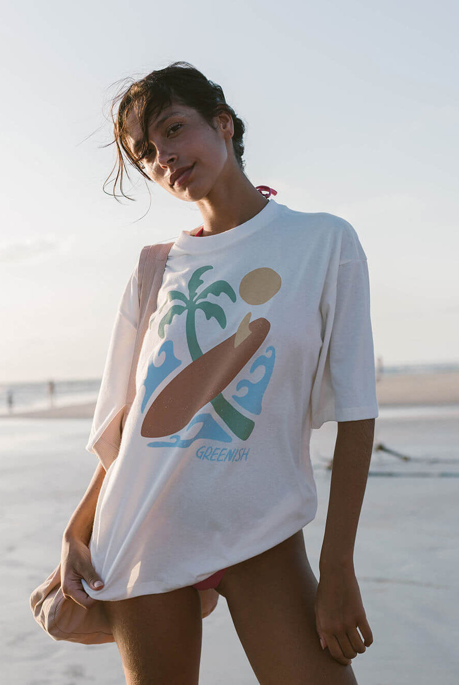 Modelo usando a camiseta oversized Sea Vibe da Greenish, na praia, com estampa tropical e estilo despojado.