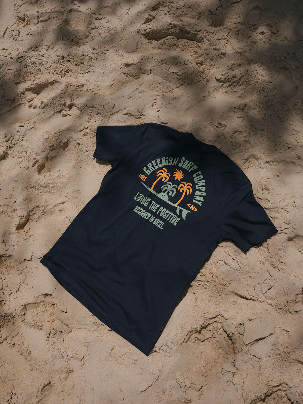 Camiseta azul marinho Greenish Living The Positive, estampada com tema tropical, sobre a areia do fundo.