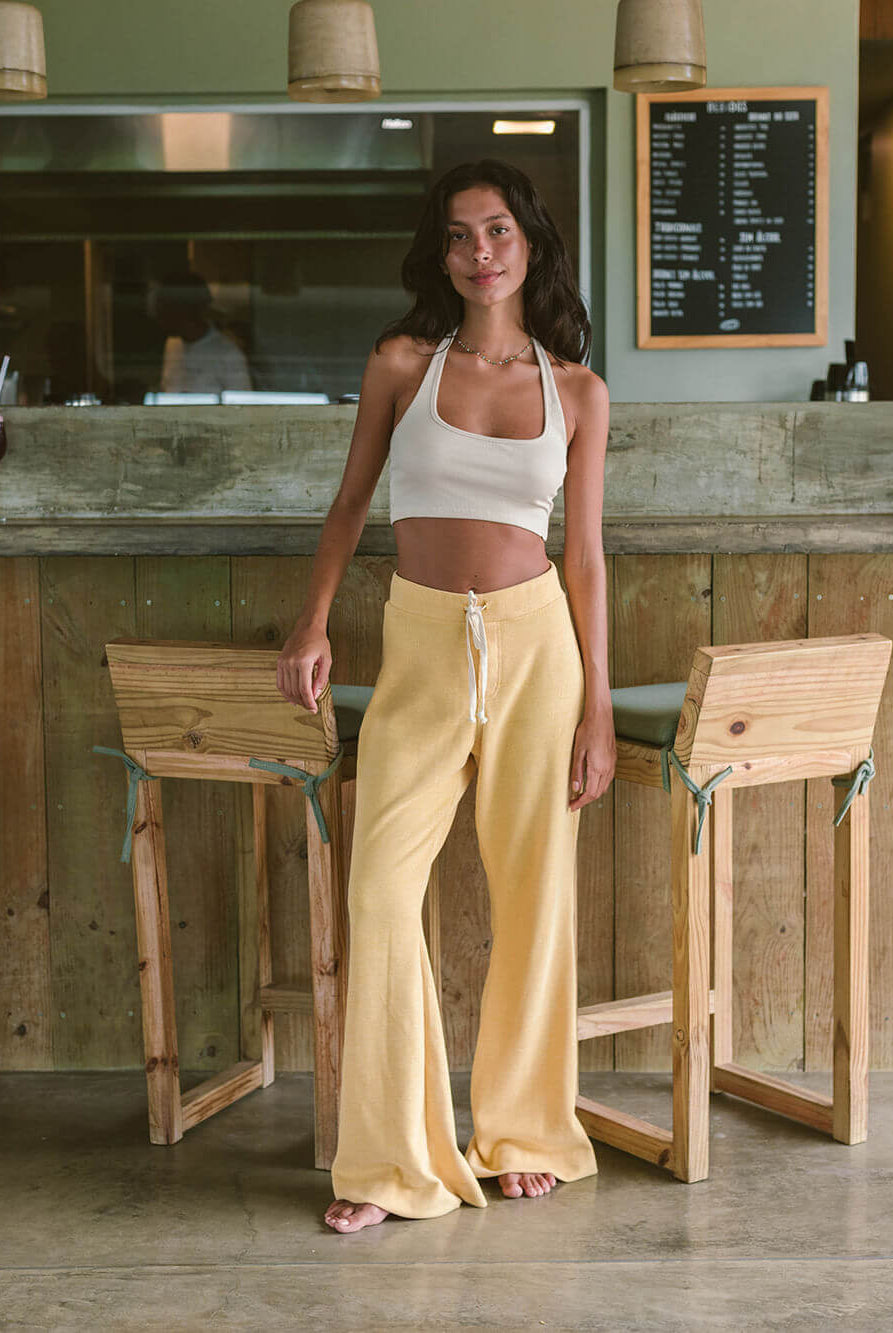 Modelo usando calça Heaven amarela da Greenish, em ambiente moderno com bar ao fundo.