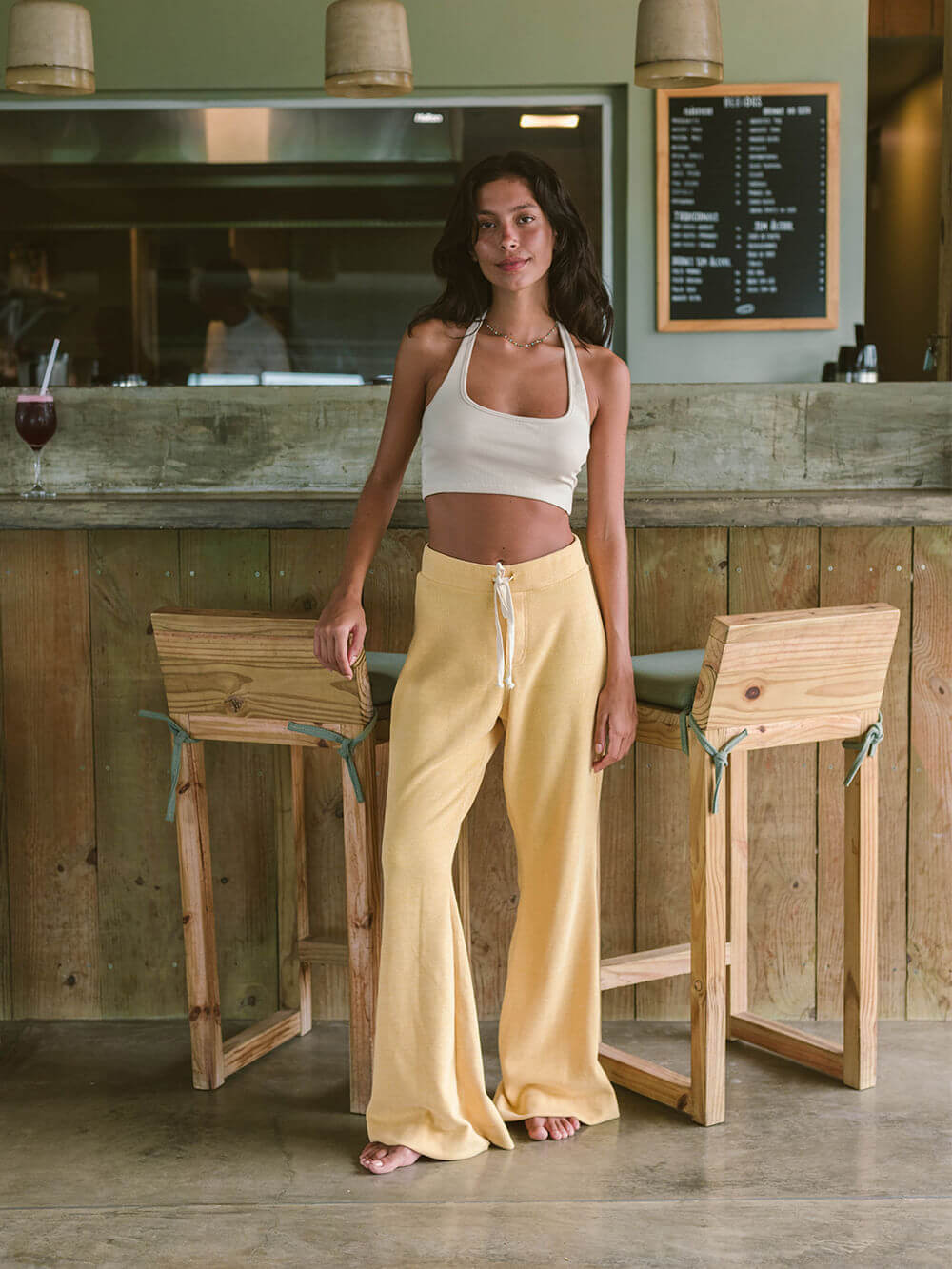 Modelo usando calça Heaven amarela da Greenish, em ambiente moderno com bar ao fundo.