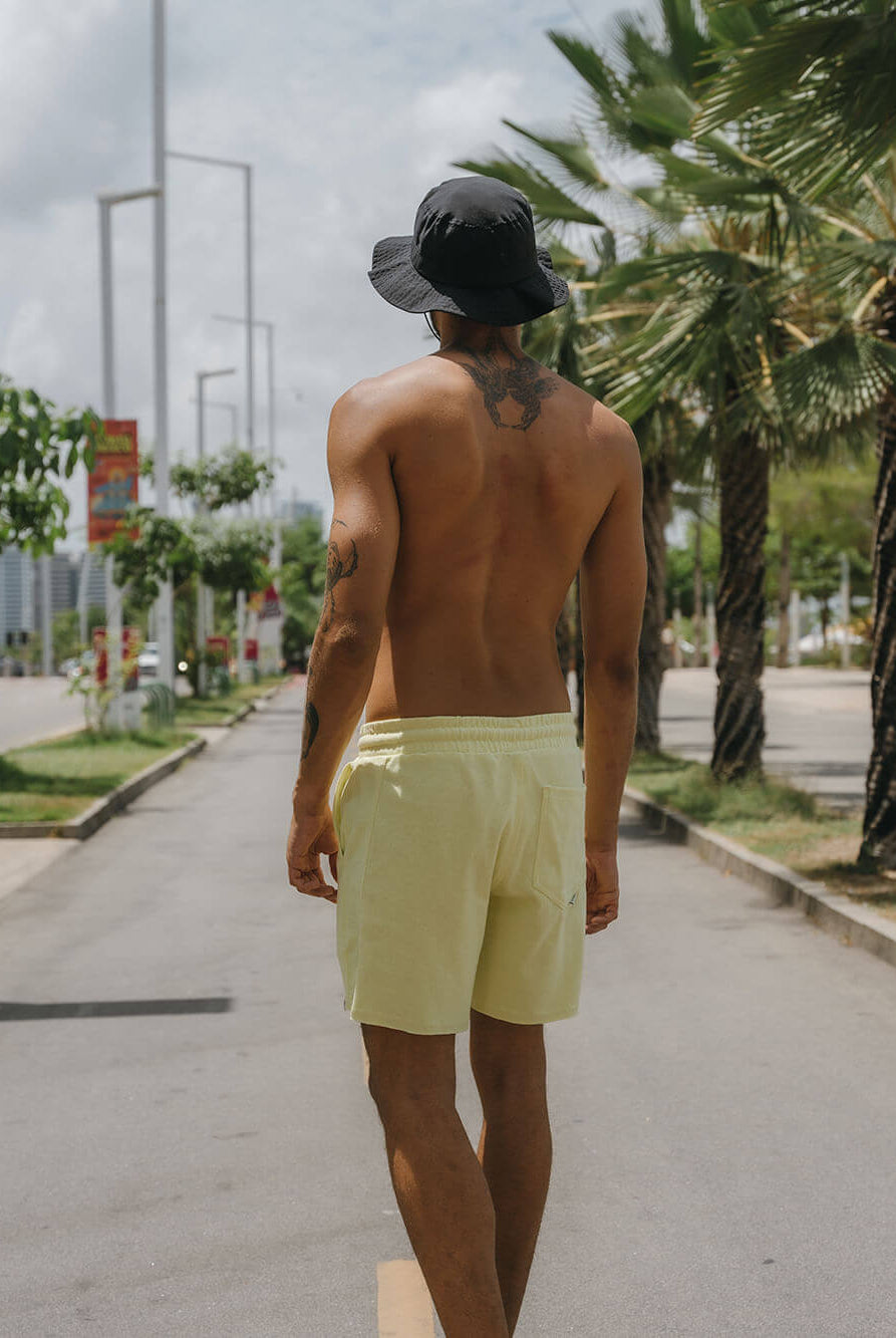 Homem com short amarelo e chapéu, caminhando em rua sob palmeiras, vibe casual com estilo Greenish.