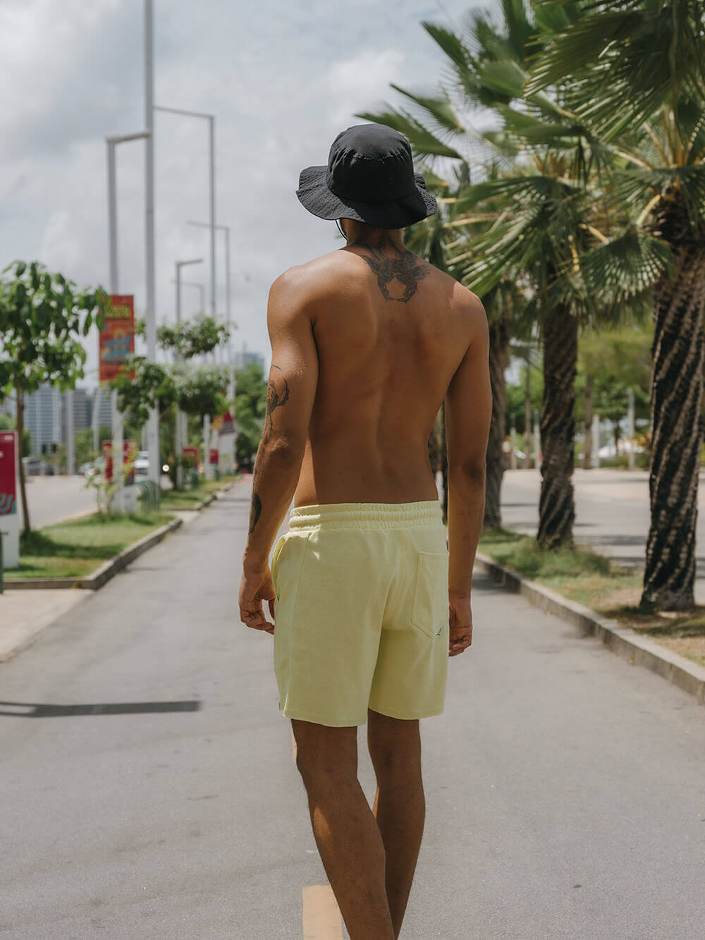 Homem com short amarelo e chapéu, caminhando em rua sob palmeiras, vibe casual com estilo Greenish.