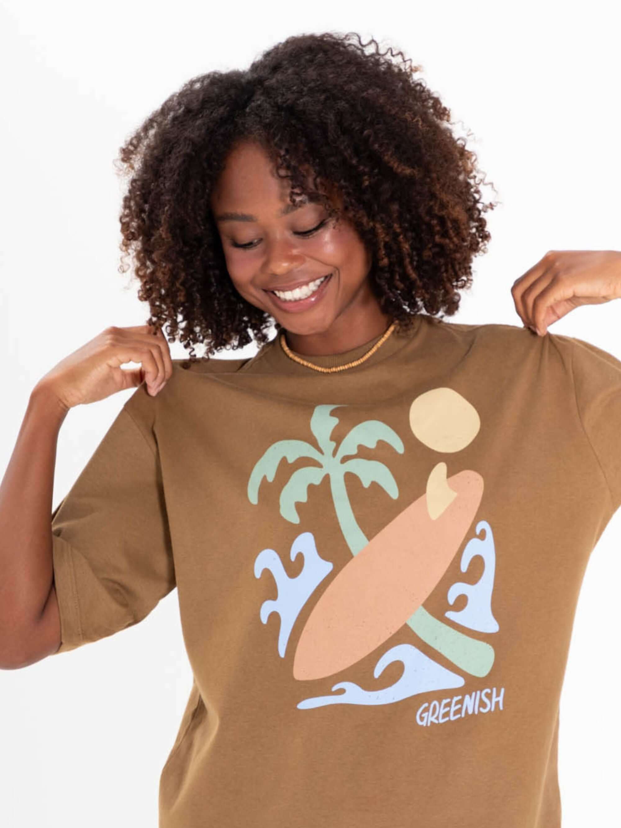 Modelo sorridente usando camiseta oversized cáqui da Greenish com estampa de surf e palmeiras.