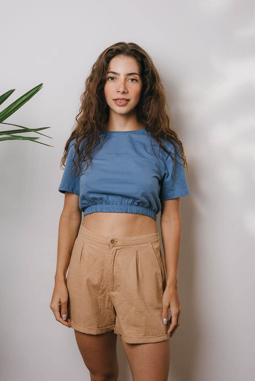 Modelo usando a Blusa Sally azul médio da Greenish, com shorts claros em um ambiente iluminado.