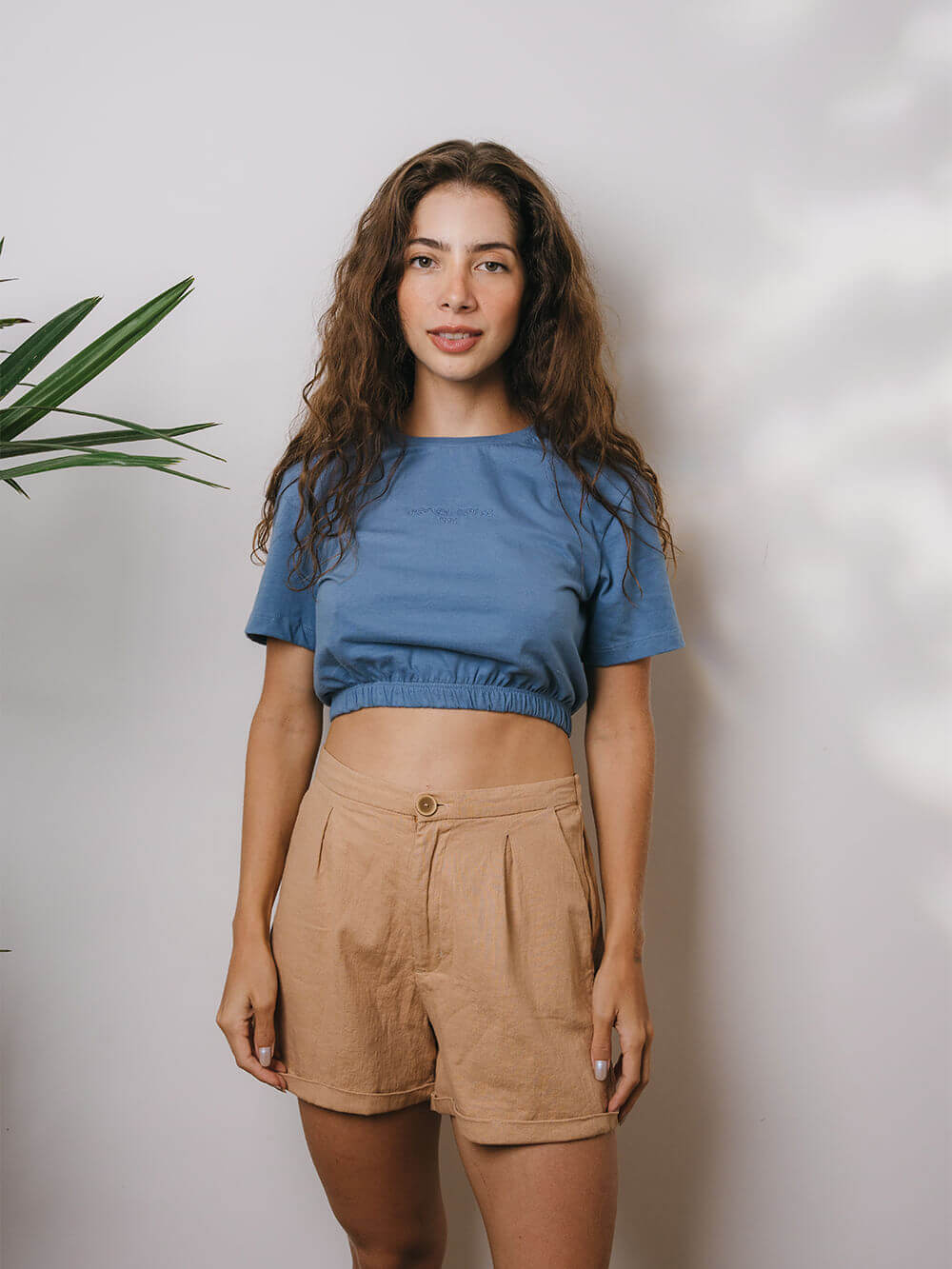 Modelo usando a Blusa Sally azul médio da Greenish, com shorts claros em um ambiente iluminado.