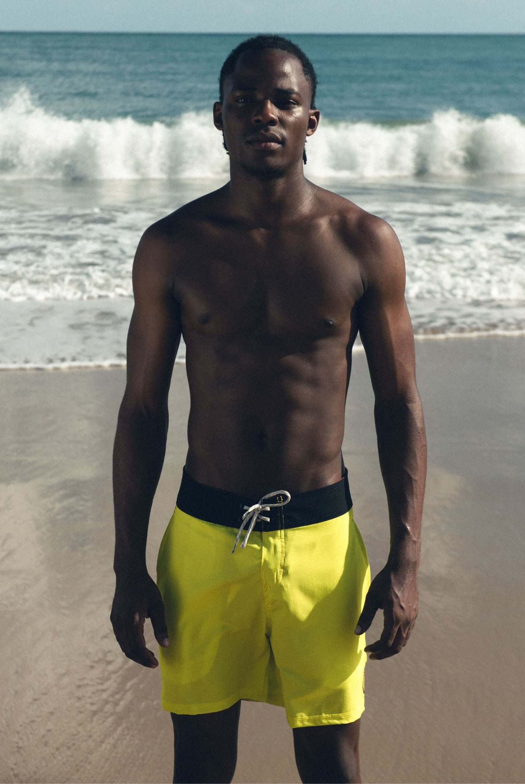 Surfista usando bermuda Boardwalk Sunny Viber Amarelo da Greenish na praia, com ondas ao fundo.