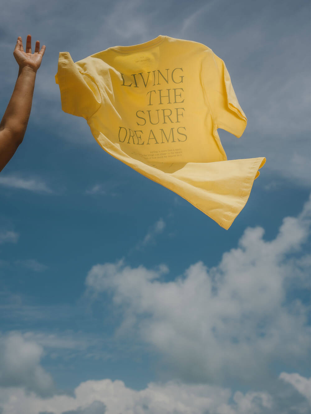 Camiseta amarela Living The Surf Dreams da marca Greenish, com céu azul ao fundo, ideal para amantes do surf.