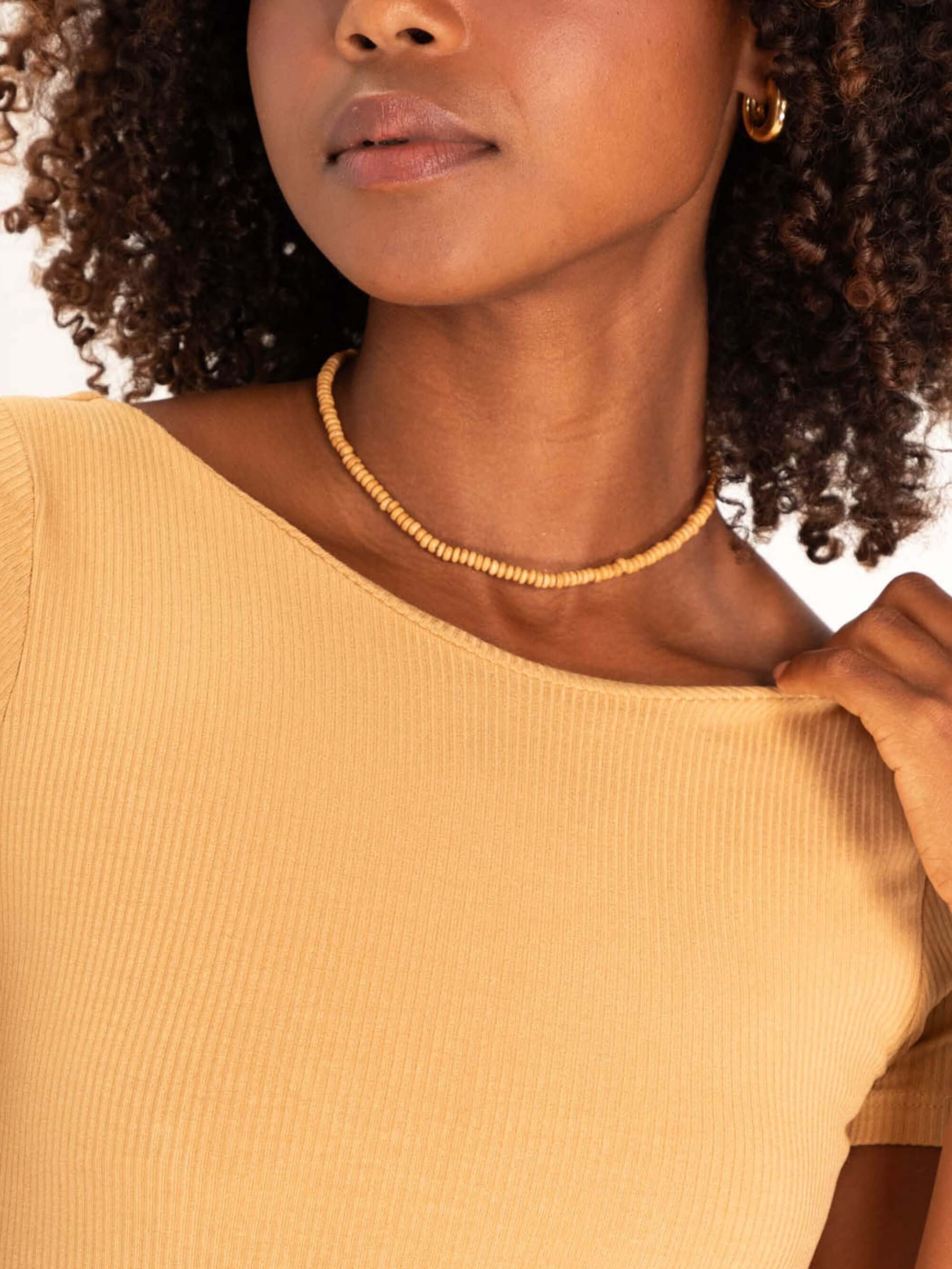 Close-up de mulher com blusa amarelo claro, destacando colar e penteado. Estilo moderno e elegante.