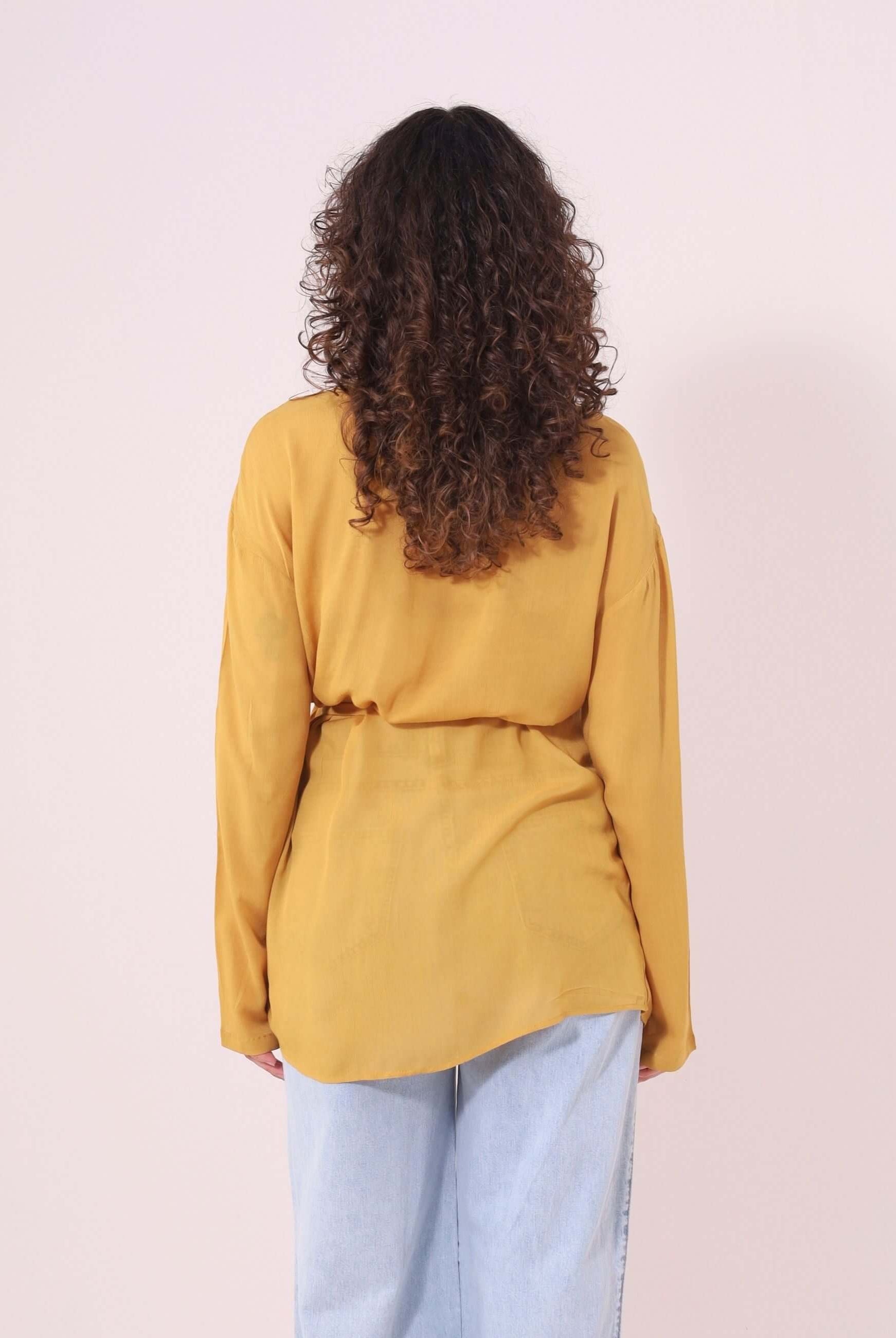 Mulher com cabelos cacheados veste Blusa Kalu Mostarda Greenish, feita de viscose com design fluido e elegante, vista de costas.