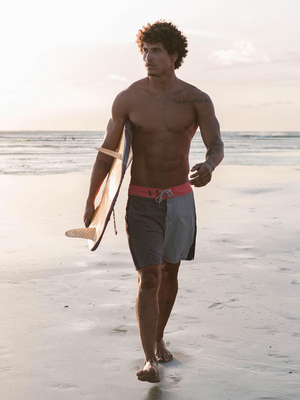 Homem surfista caminhando na praia com prancha, usando boardshorts Greenish, destacando estilo e conforto.