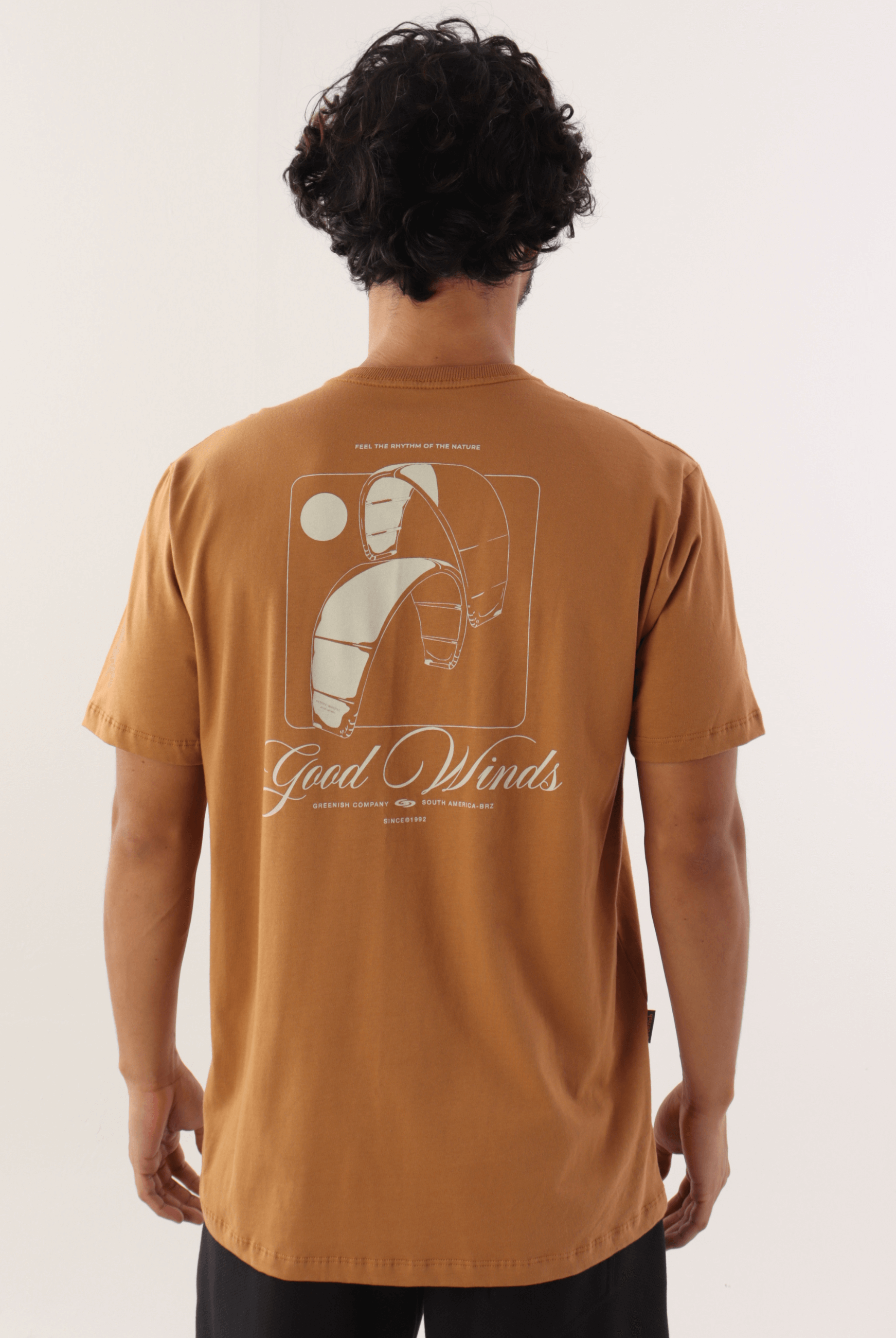 Homem usando camiseta marron da marca Greenish com estampa "Kites & Good Winds" nas costas, em estilo casual e esportivo.