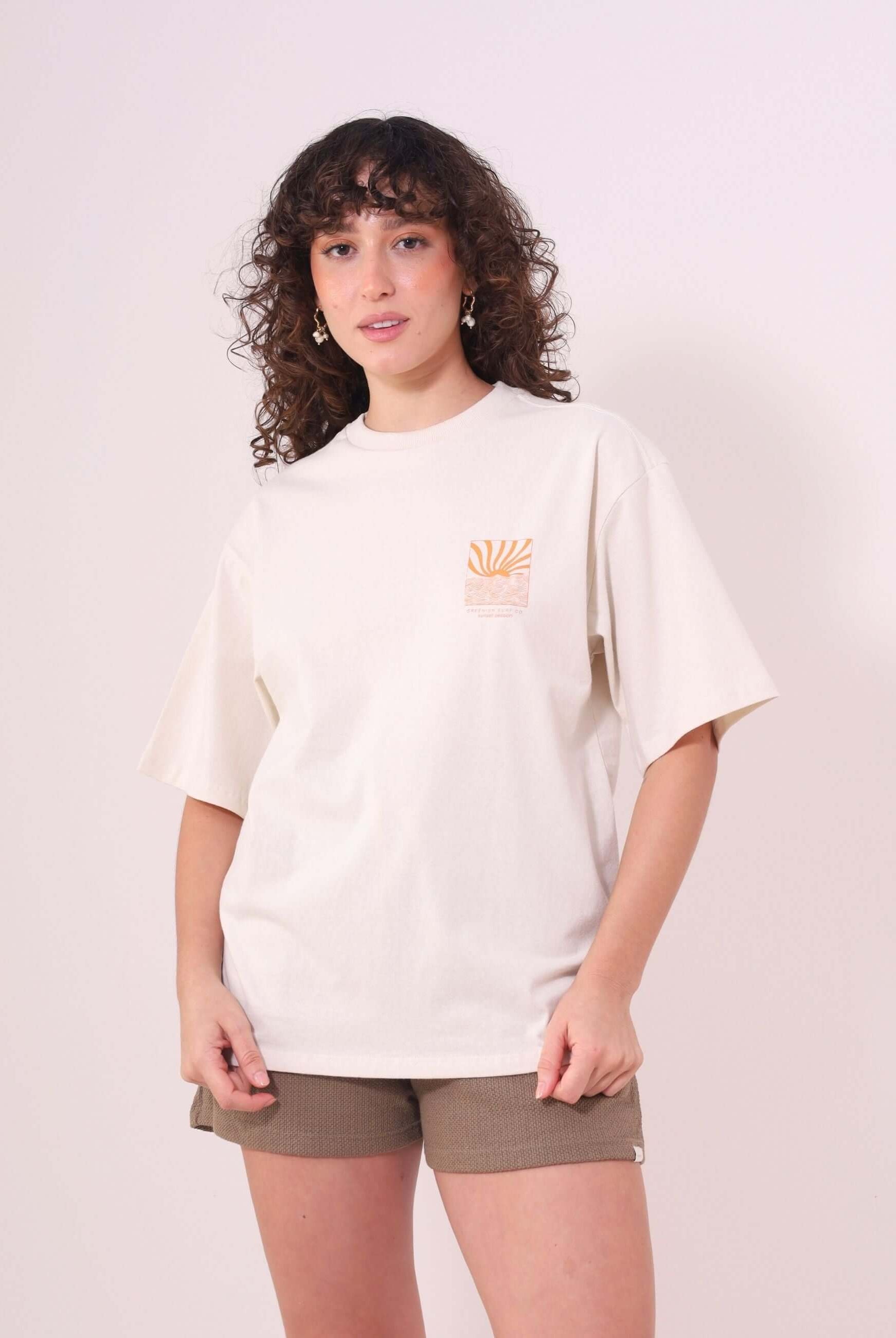 Mulher vestindo Camiseta Oversized Sunset Session da Greenish, cor natural, estampa de ondas e pôr do sol, feita de algodão macio.