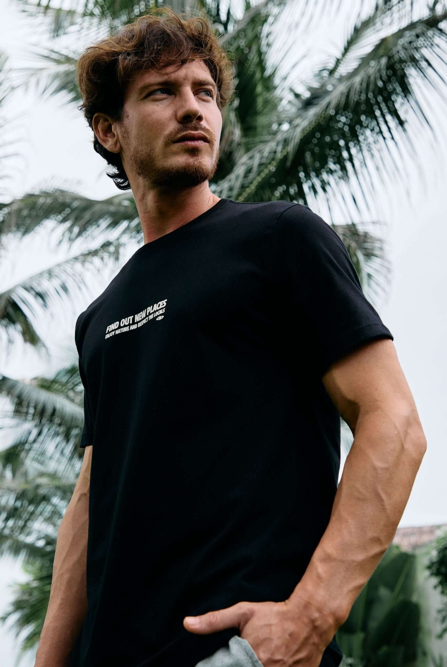 Homem usando camiseta preta Seagull Greenish ao ar livre com palmeiras ao fundo, evocando liberdade e natureza. 100% algodão.