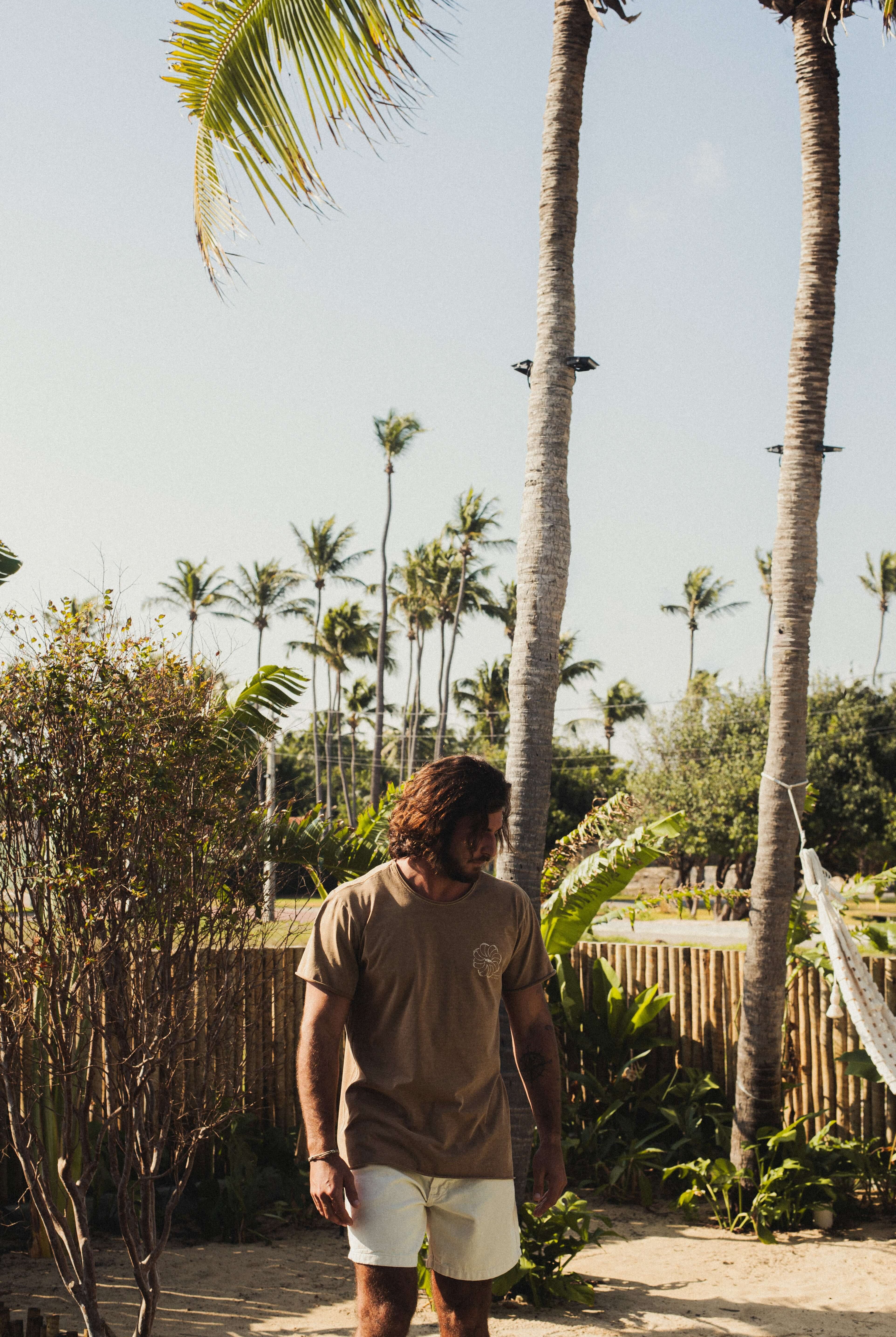 Homem vestindo Camiseta Stone Feel The Winds - Marrom da Greenish, em um cenário tropical. Toque macio e visual rústico. 100% algodão.