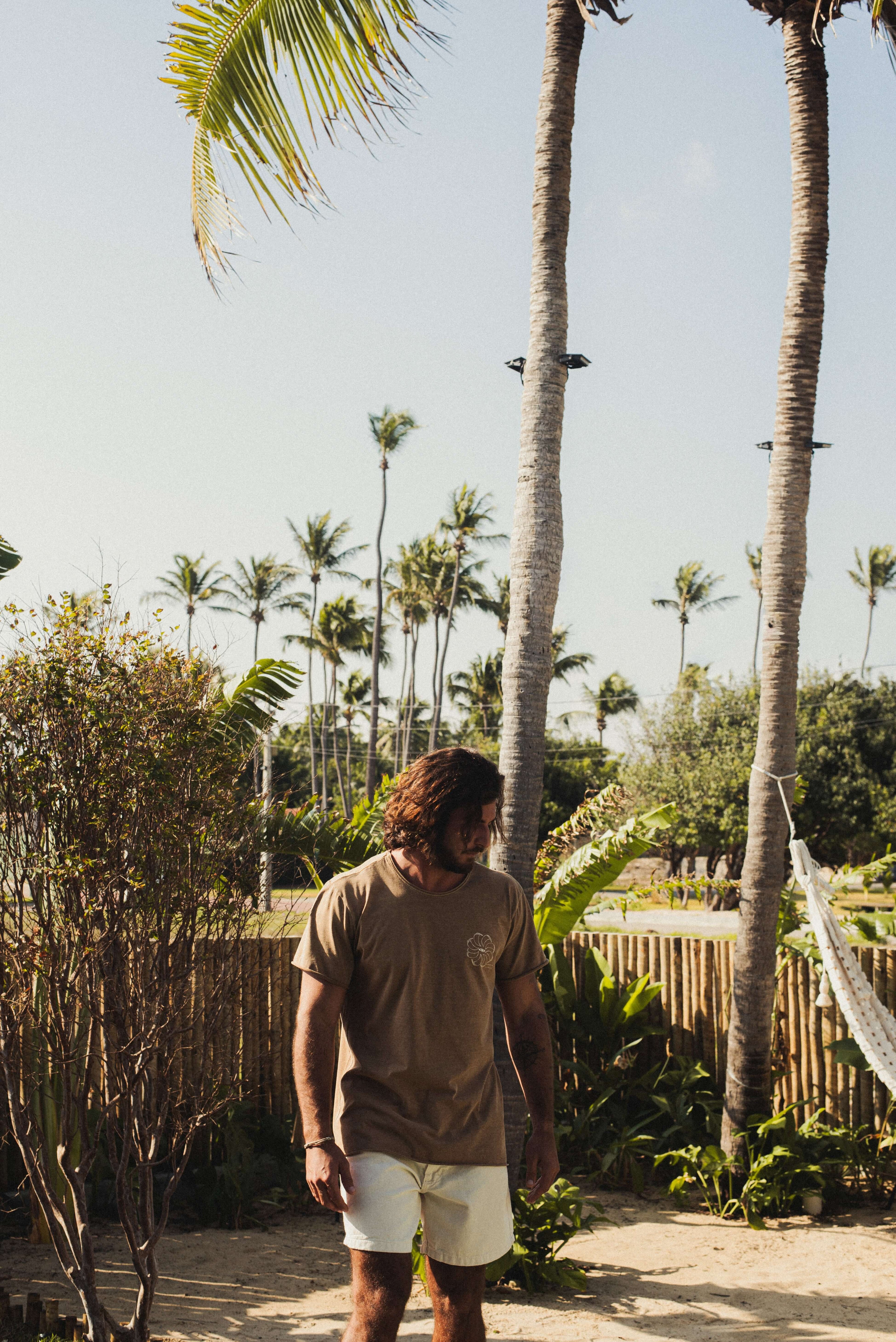 Homem vestindo Camiseta Stone Feel The Winds - Marrom da Greenish, em um cenário tropical. Toque macio e visual rústico. 100% algodão.