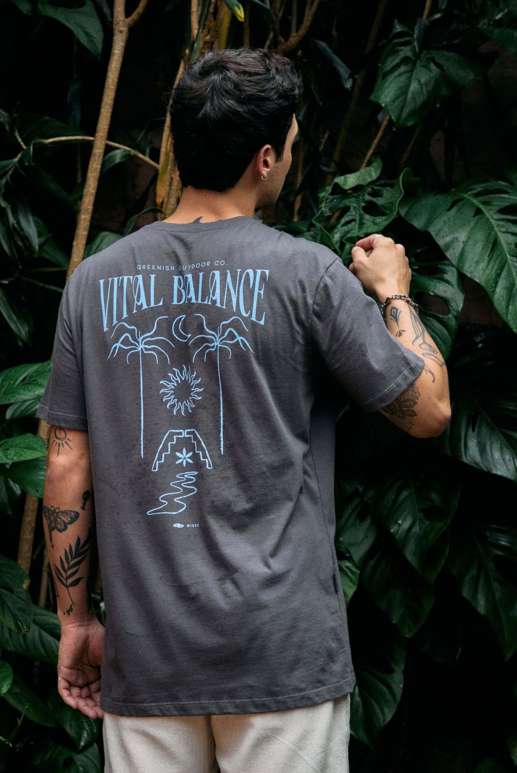 Homem vestindo camiseta Greenish 'The Seed, The Sun and Moon' grafite, com estampa de equilíbrio natural, em cenário de plantas tropicais.