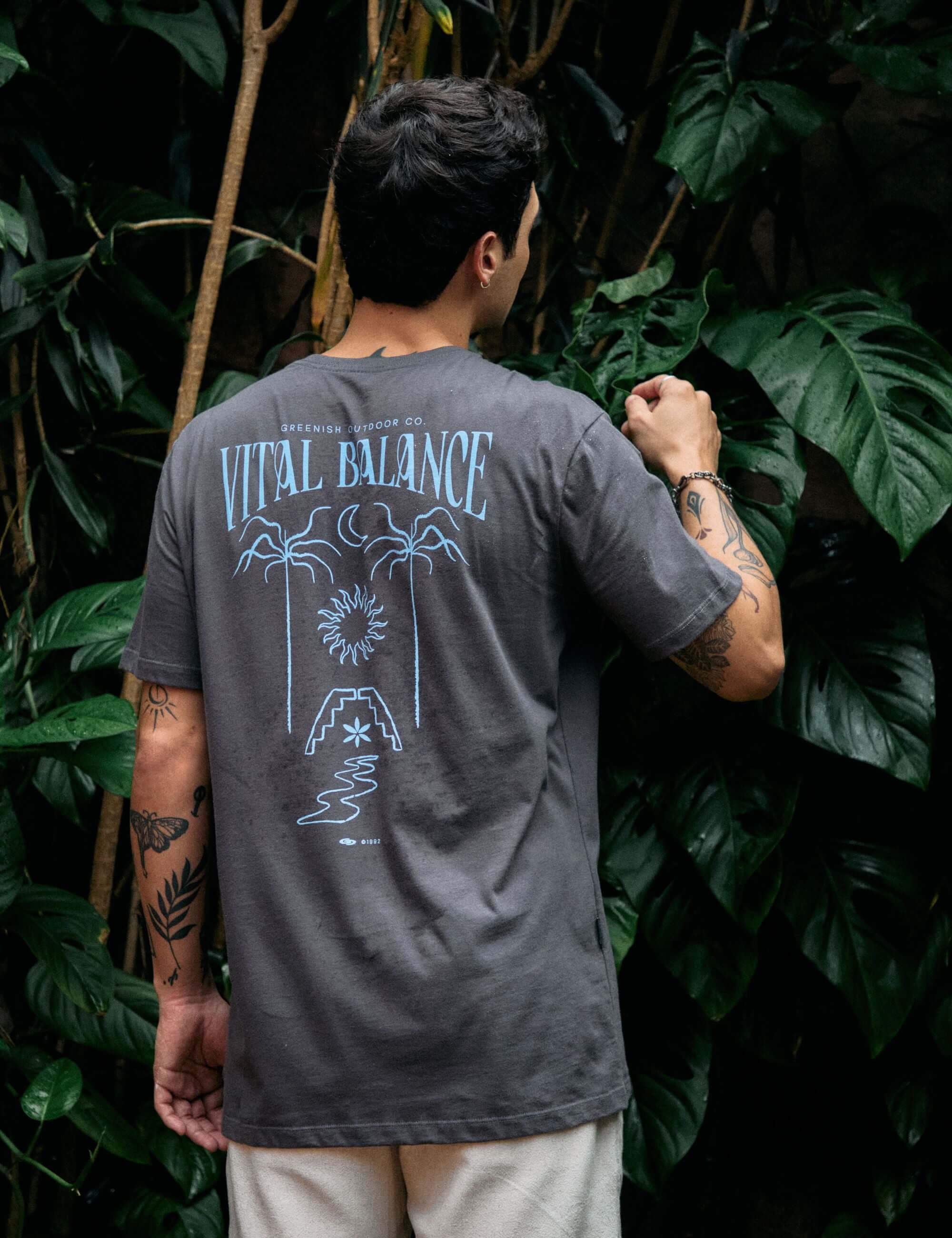 Homem vestindo camiseta Greenish 'The Seed, The Sun and Moon' grafite, com estampa de equilíbrio natural, em cenário de plantas tropicais.
