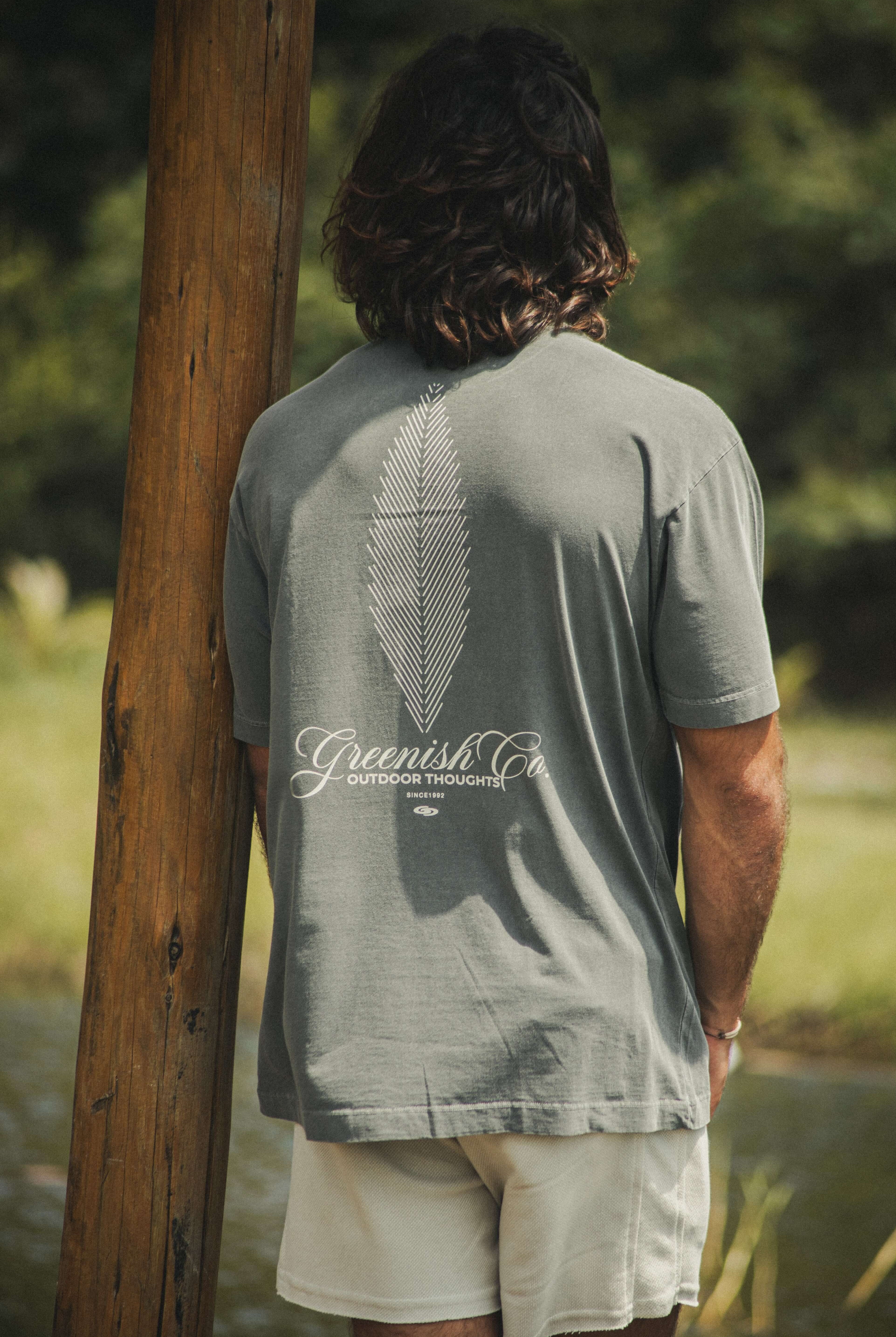 Homem vestindo a Camiseta Oversized Stong Leaf Plank - Chumbo da Greenish, com estampa de folha e logo, em um ambiente natural.