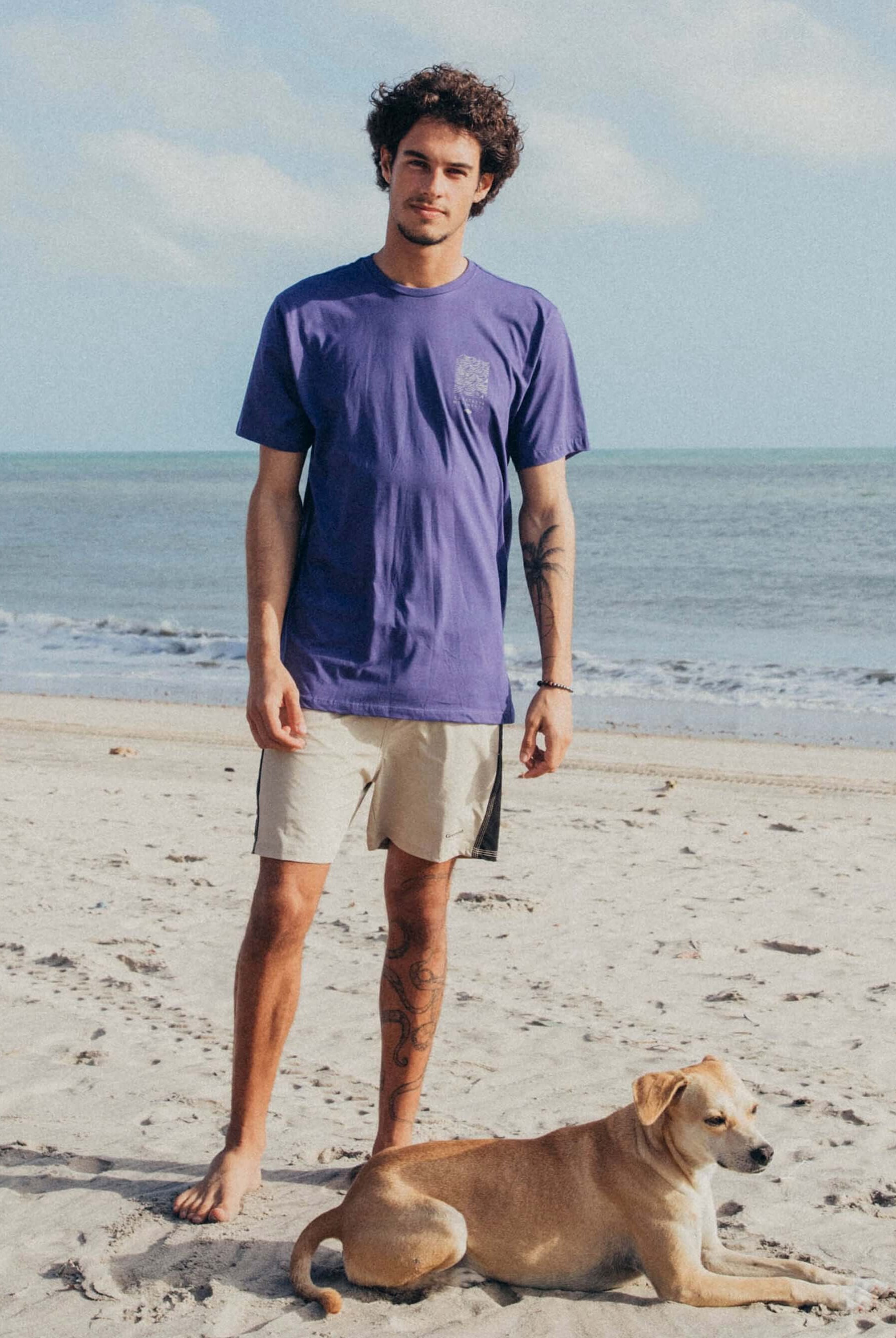Homem na praia usando camiseta roxa da Greenish e short claro, com cachorro ao seu lado.