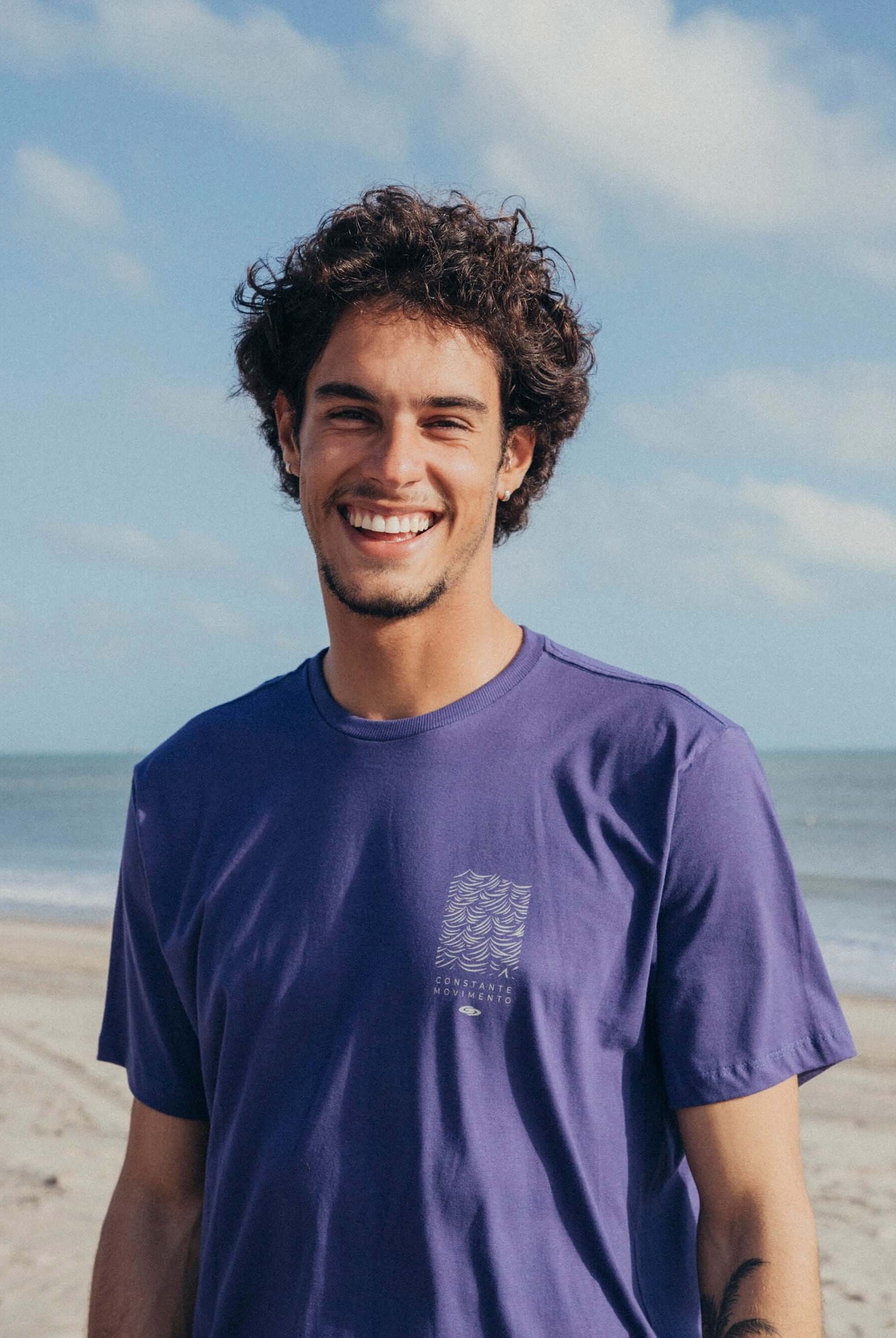 Camiseta Constantemente Movimento azul royal da marca Greenish, modelo em foto, à beira da praia.