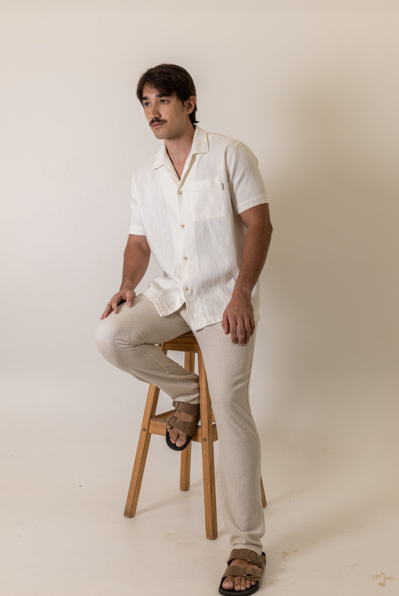 Man wearing Greenish Camisa Botao High Tide - Off White sitting on stool in light background, showcasing casual style.