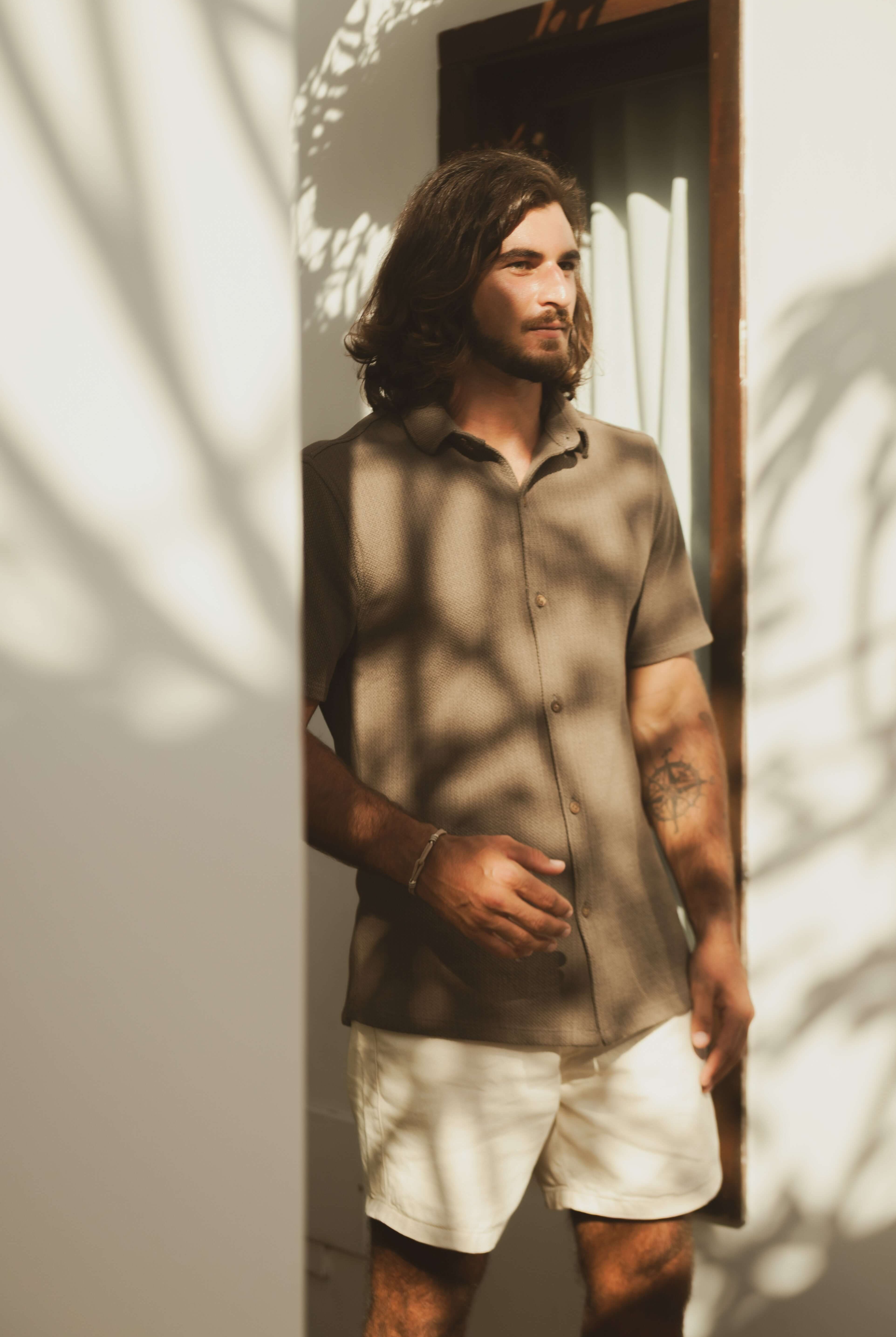 Homem usando Camisa Botão Maré - Verde Escuro, destacando elegância casual e conforto em um ambiente iluminado por sombras de folhas.
