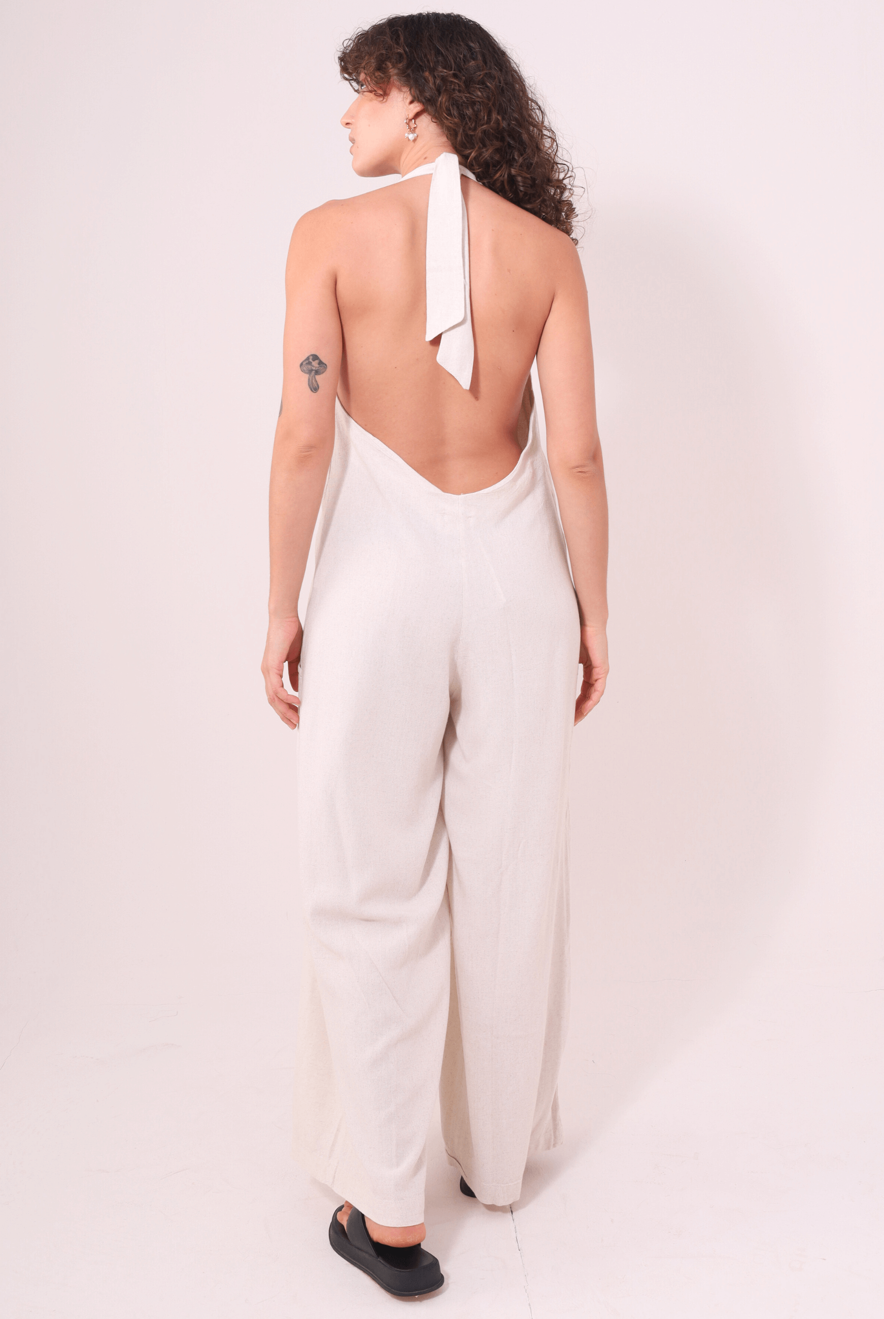 Mulher vestindo Macacão Longo Gaia - Natural por Greenish, de costas, destacando decote halter, pernas amplas e amarração no pescoço.