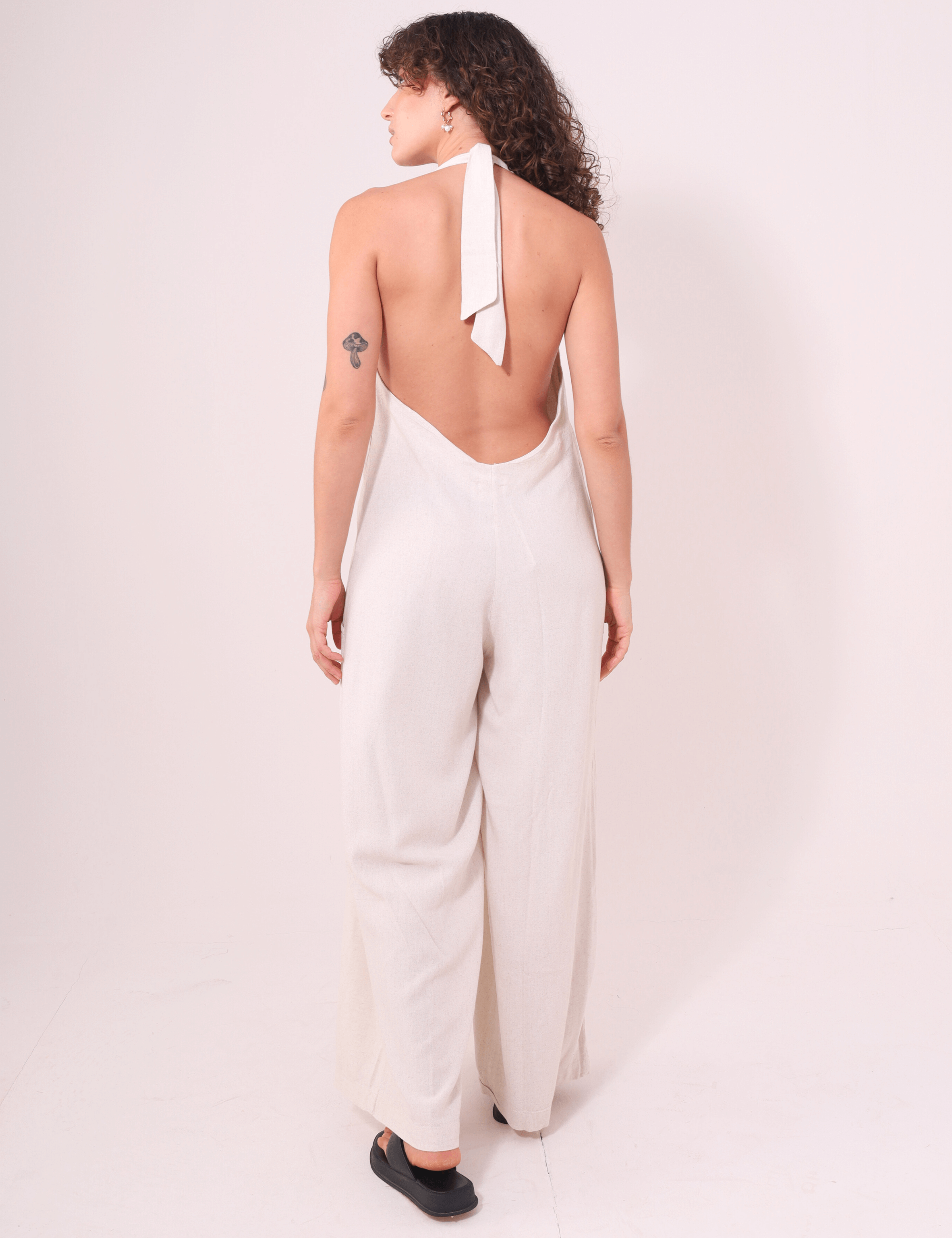 Mulher vestindo Macacão Longo Gaia - Natural por Greenish, de costas, destacando decote halter, pernas amplas e amarração no pescoço.