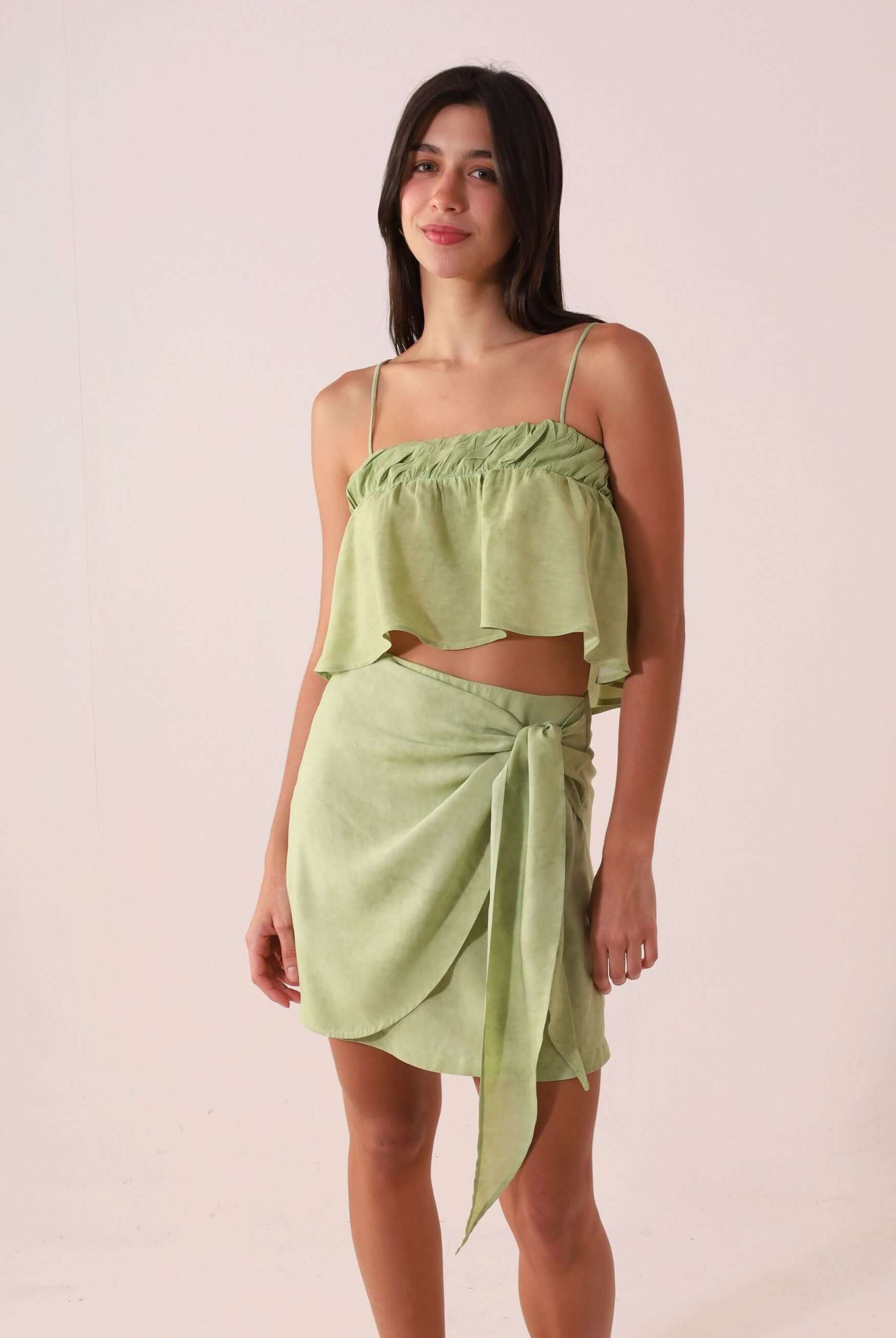 Modelo usa a SAIA CURTA THÁ - VERDE por Greenish, destacando amarração lateral e sobreposição. Saia em viscose macia e leve.