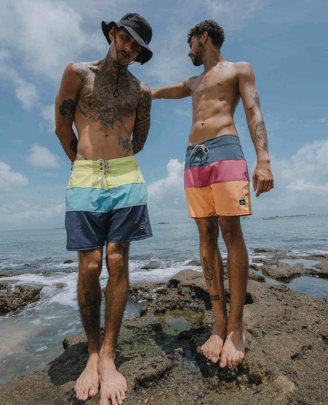 Dois homens na praia usando bermudas de banho coloridas da Greenish, destacando o estilo e conforto do Boardwalk Three Panels.