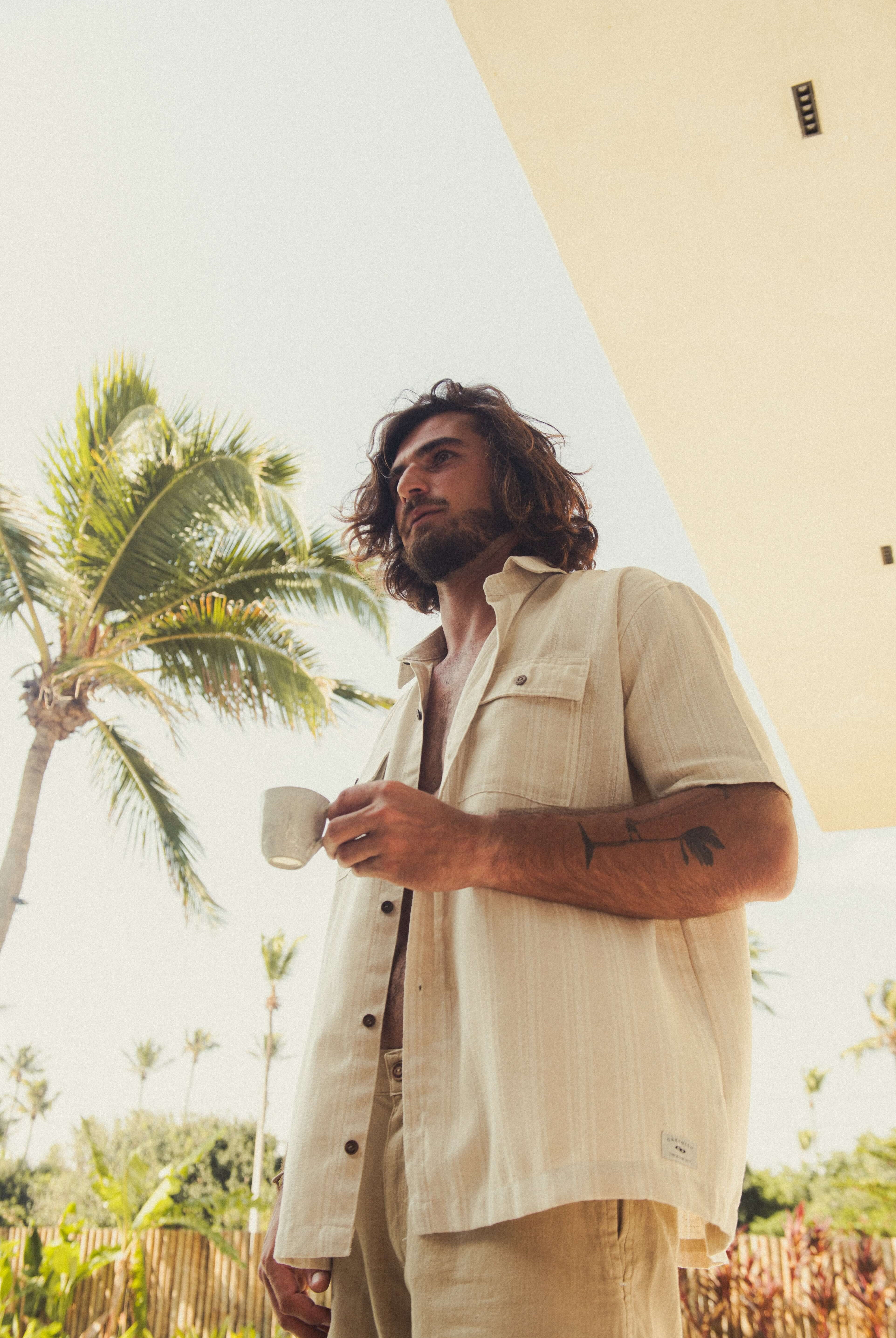 Homem com Camisa Botão Linho Listrado Oversized - Natural da marca Greenish, em cenário tropical ao ar livre. Moda utilitária sofisticada.