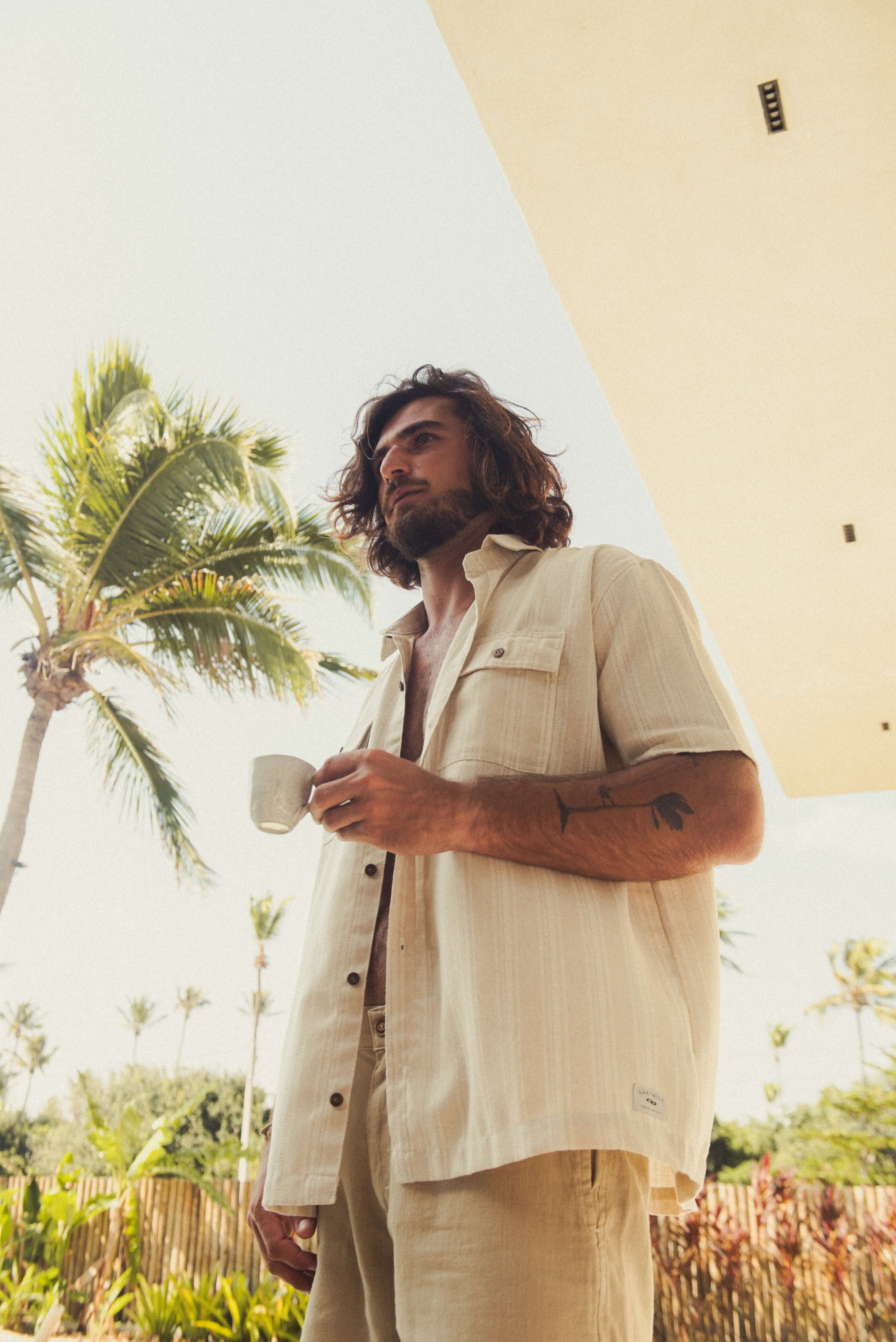 Homem com Camisa Botão Linho Listrado Oversized - Natural da marca Greenish, em cenário tropical ao ar livre. Moda utilitária sofisticada.