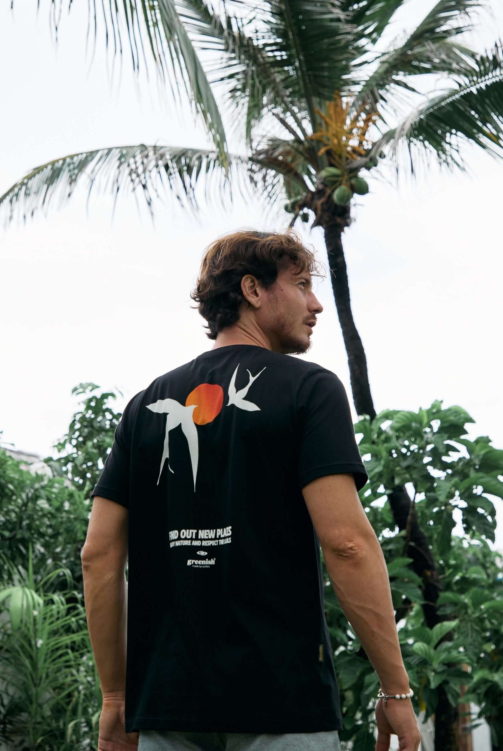 Homem vestindo a camiseta Seagull preta da Greenish, inspirada na natureza, com arte de gaivota e sol, em frente a coqueiro e vegetação.