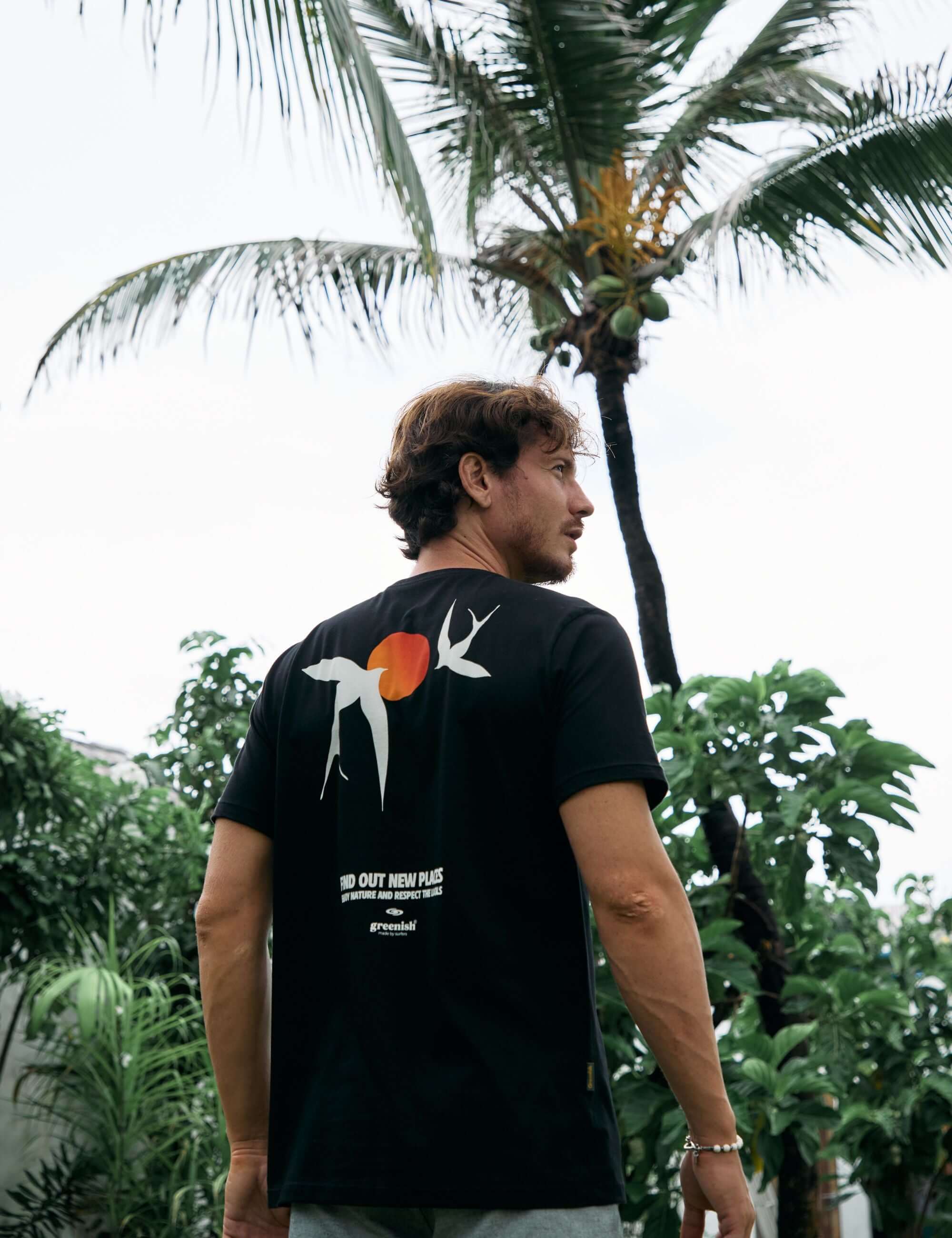 Homem vestindo a camiseta Seagull preta da Greenish, inspirada na natureza, com arte de gaivota e sol, em frente a coqueiro e vegetação.