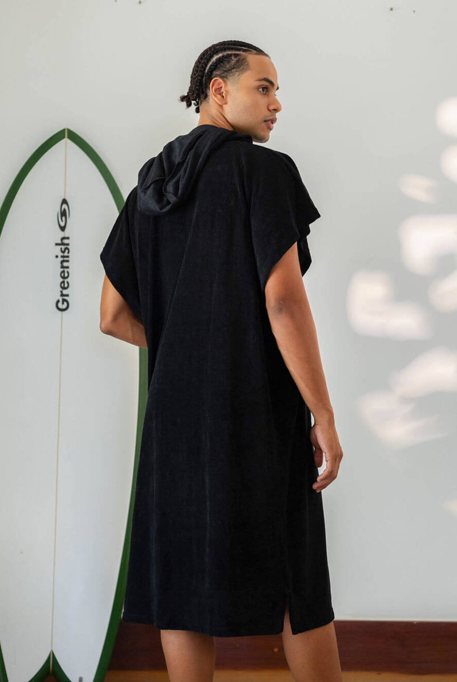 Homem usando poncho adulto preto Greenish, com capuz e surfboard ao fundo, ideal para praia e esportes aquáticos.