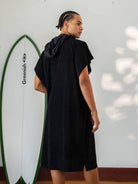 Homem usando poncho adulto preto Greenish, com capuz e surfboard ao fundo, ideal para praia e esportes aquáticos.