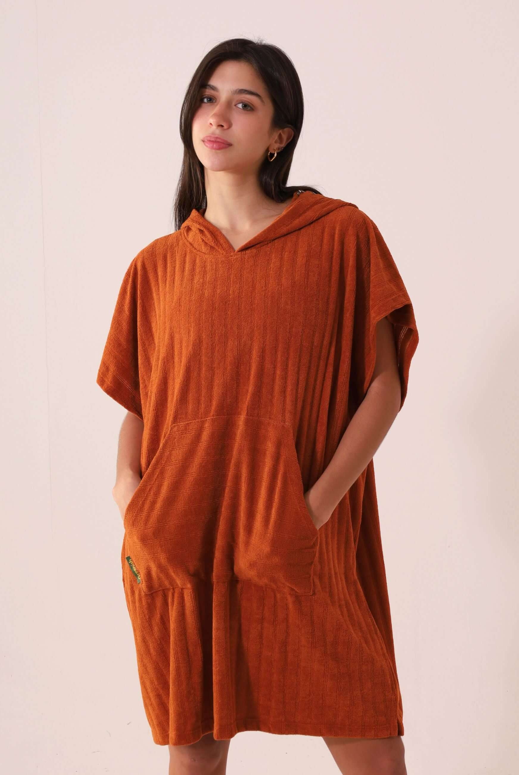 Mulher vestindo o PONCHO CURTO CANELADO - CARAMELO da Greenish, em tecido atoalhado com capuz e bolsos canguru, ideal para a praia.