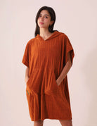 Mulher vestindo o PONCHO CURTO CANELADO - CARAMELO da Greenish, em tecido atoalhado com capuz e bolsos canguru, ideal para a praia.