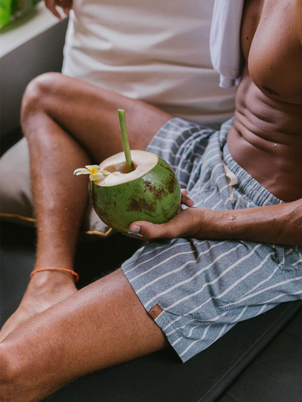 Homem relaxando em sofá com coco verde na mão, usando short listrado da marca Greenish.
