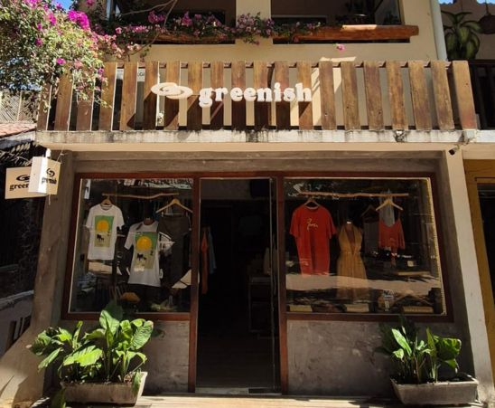 Fachada da loja 'greenish' com roupas expostas e plantas na entrada, em ambiente ensolarado.