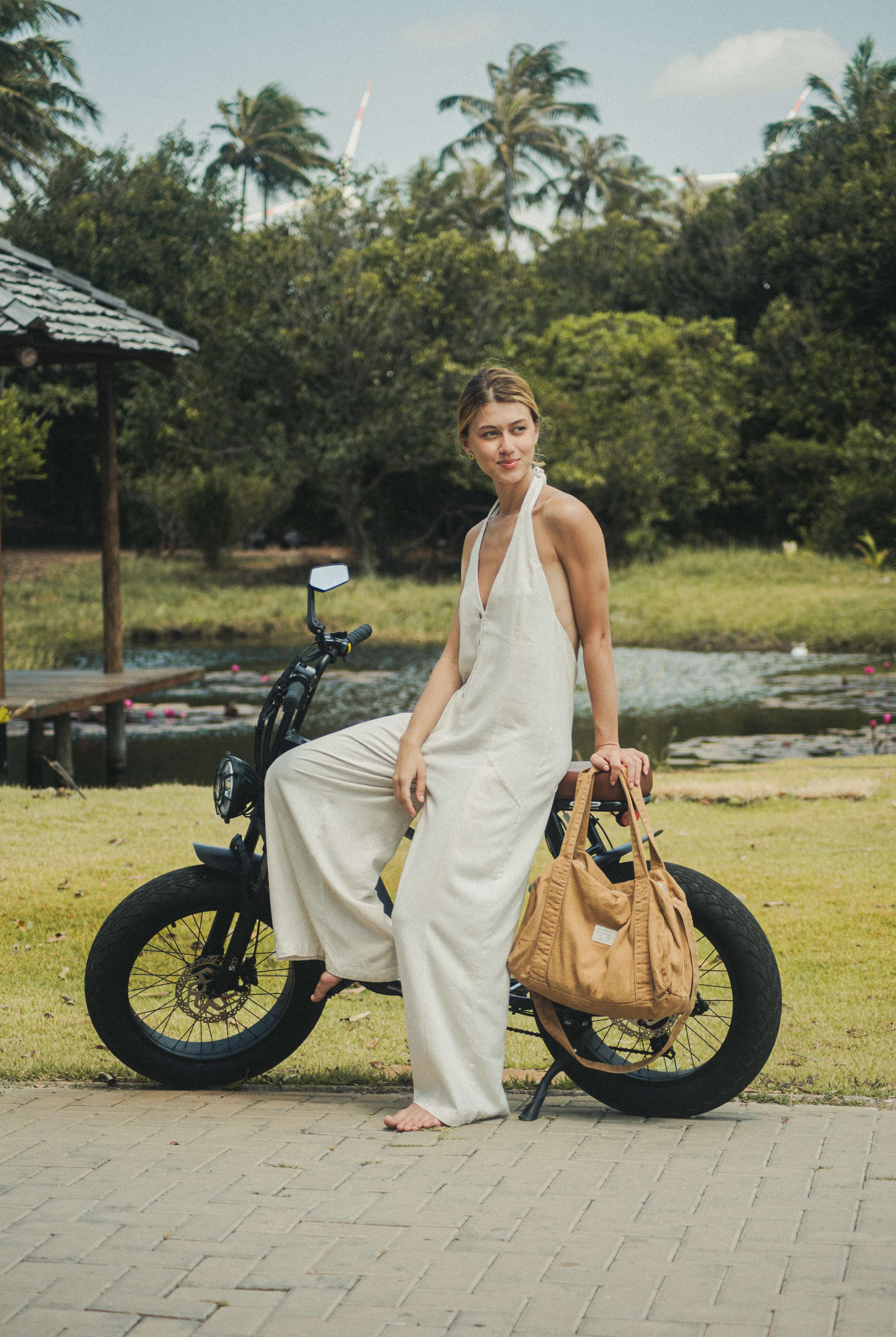 Mulher usando o Macacão Longo Gaia - Natural da Greenish, tecido de linho leve e fresco, em ambiente natural ao lado de uma bicicleta elétrica.