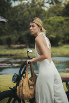 Mulher usando Macacão Longo Gaia - Natural da Greenish, em linho leve, com decote halter e amarração, ao lado de uma bicicleta em ambiente natural.