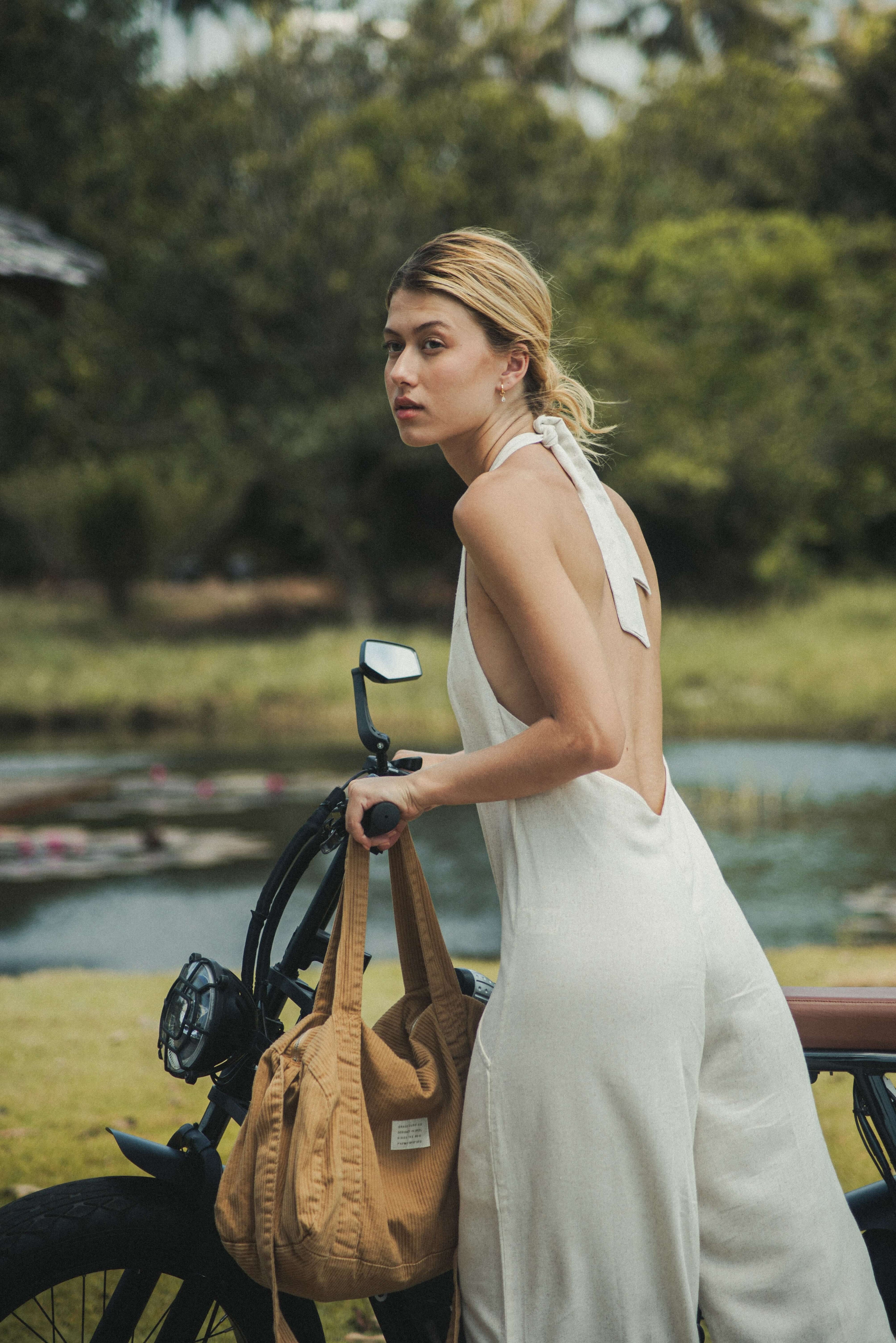 Mulher usando Macacão Longo Gaia - Natural da Greenish, em linho leve, com decote halter e amarração, ao lado de uma bicicleta em ambiente natural.