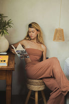 Mulher vestindo Macacão Longo Riviera - Cacau da Greenish, sem alças e elegante, leitura de revista em ambiente aconchegante.