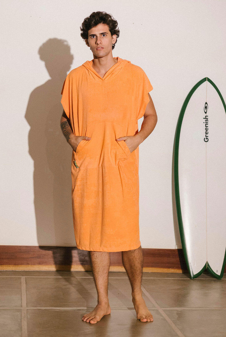 Homem usando poncho adulto bolso canguru coral da Greenish, ao lado de uma prancha de surf.