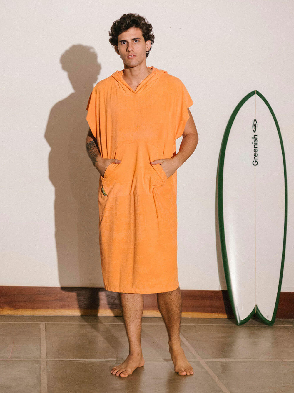 Homem usando poncho adulto bolso canguru coral da Greenish, ao lado de uma prancha de surf.