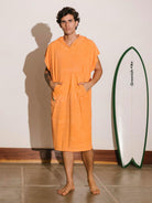 Homem usando poncho adulto bolso canguru coral da Greenish, ao lado de uma prancha de surf.