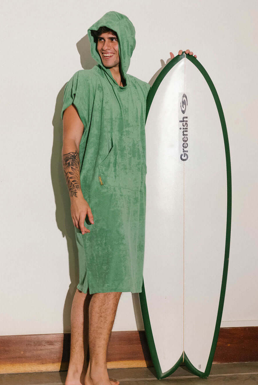Homem usando poncho adulto Greenish verde, segurando uma prancha de surf, perfeito para praia e esportes aquáticos.