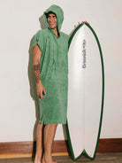 Homem usando poncho adulto Greenish verde, segurando uma prancha de surf, perfeito para praia e esportes aquáticos.